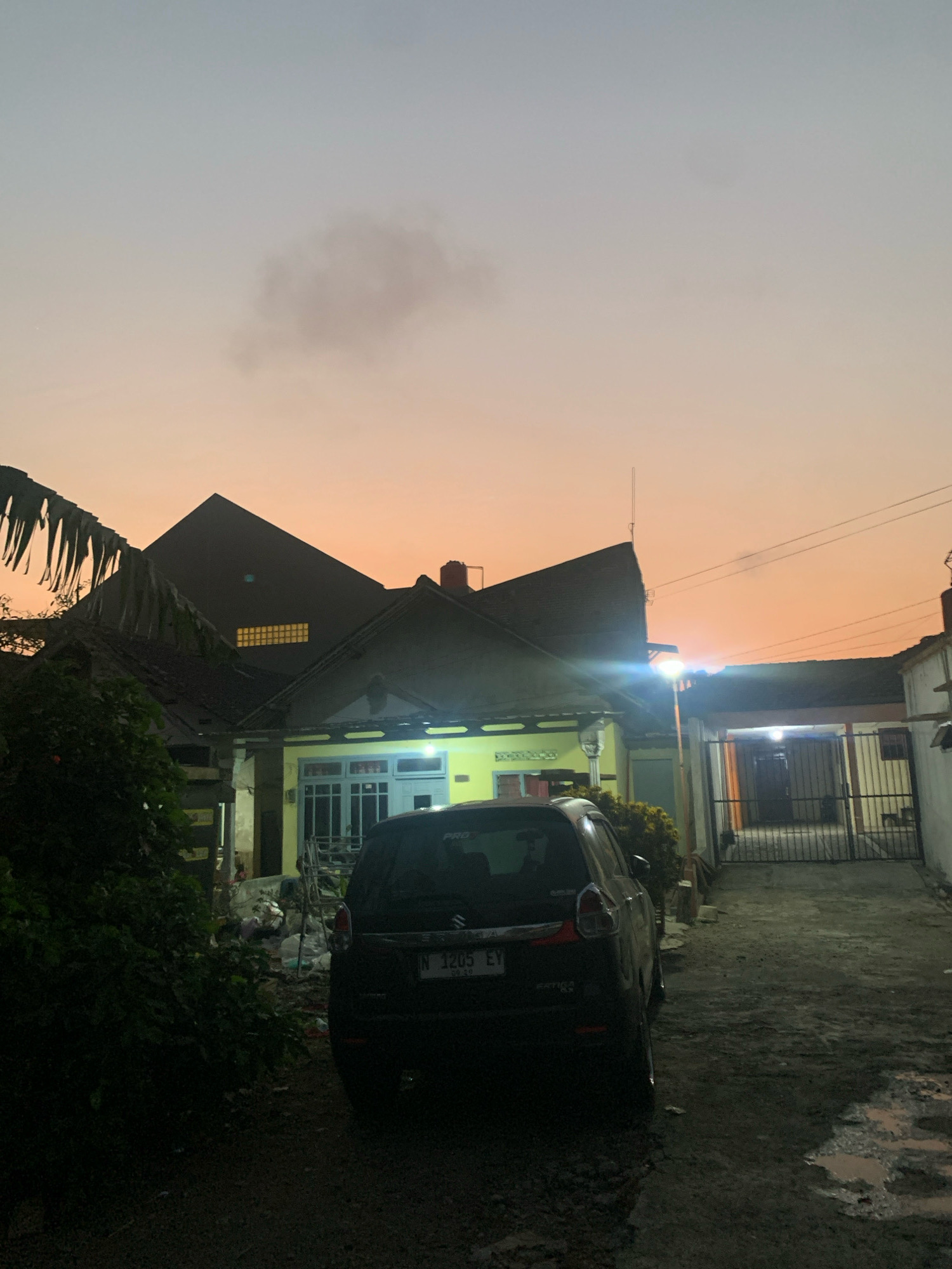 The image shows a residential street scene in Bululawang, Indonesia, during twilight. The sky is a gradient of soft orange and pale blue, with a few wispy clouds. In the foreground, a dark-colored car is parked on a dirt or gravel driveway. To the left of the car, dense green foliage obscures part of the view.
Behind the car, a single-story house with a yellow facade and a dark-tiled roof is visible. The house has several windows, some of which are illuminated. A large, dark, angular structure, possibly another part of the house or a neighboring building, looms behind the yellow house, featuring a series of small, illuminated rectangular windows. To the right of the yellow house, a metal gate leads into a courtyard or driveway, with another building in the background. A tall, thin light post stands to the right of the yellow house, emitting a bright light. The ground appears to be a mix of dark soil and puddles of water, suggesting recent rain. The overall impression is one of a quiet, domestic scene at dusk.