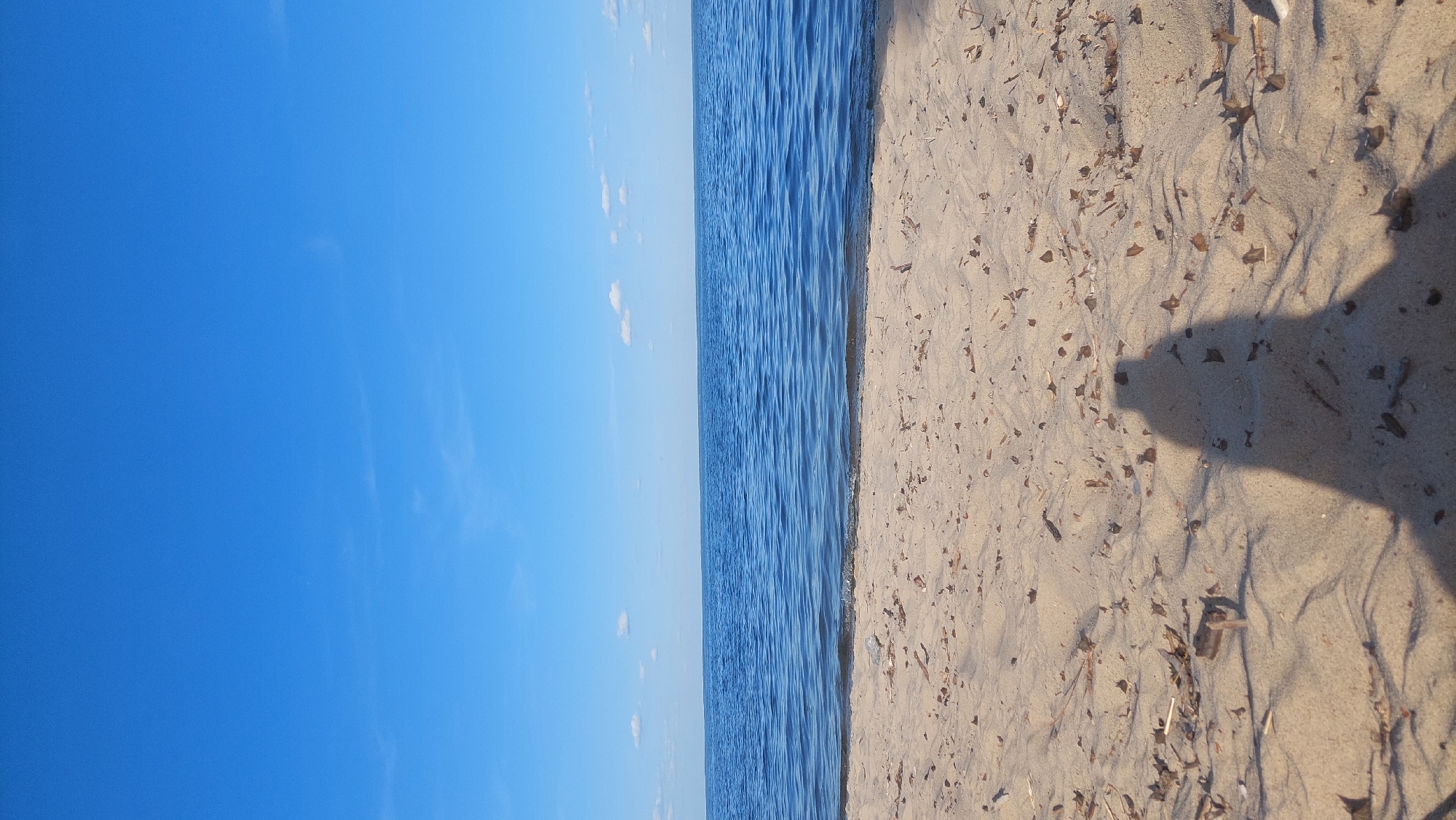 The image shows a tranquil scene at the beach in Kozarovychi, Ukraine. The foreground is dominated by light-colored sand, strewn with small pieces of dried leaves and twigs. A distinct shadow of a person, presumably the photographer, is cast on the sand, extending from the bottom right corner towards the center. Beyond the beach, a wide expanse of calm blue water stretches towards the horizon. The water reflects the clear blue sky above, which is dotted with a few scattered, small white clouds. The horizon line is sharply defined, separating the water from the sky. The overall impression is one of a peaceful, sunny day at the seaside.