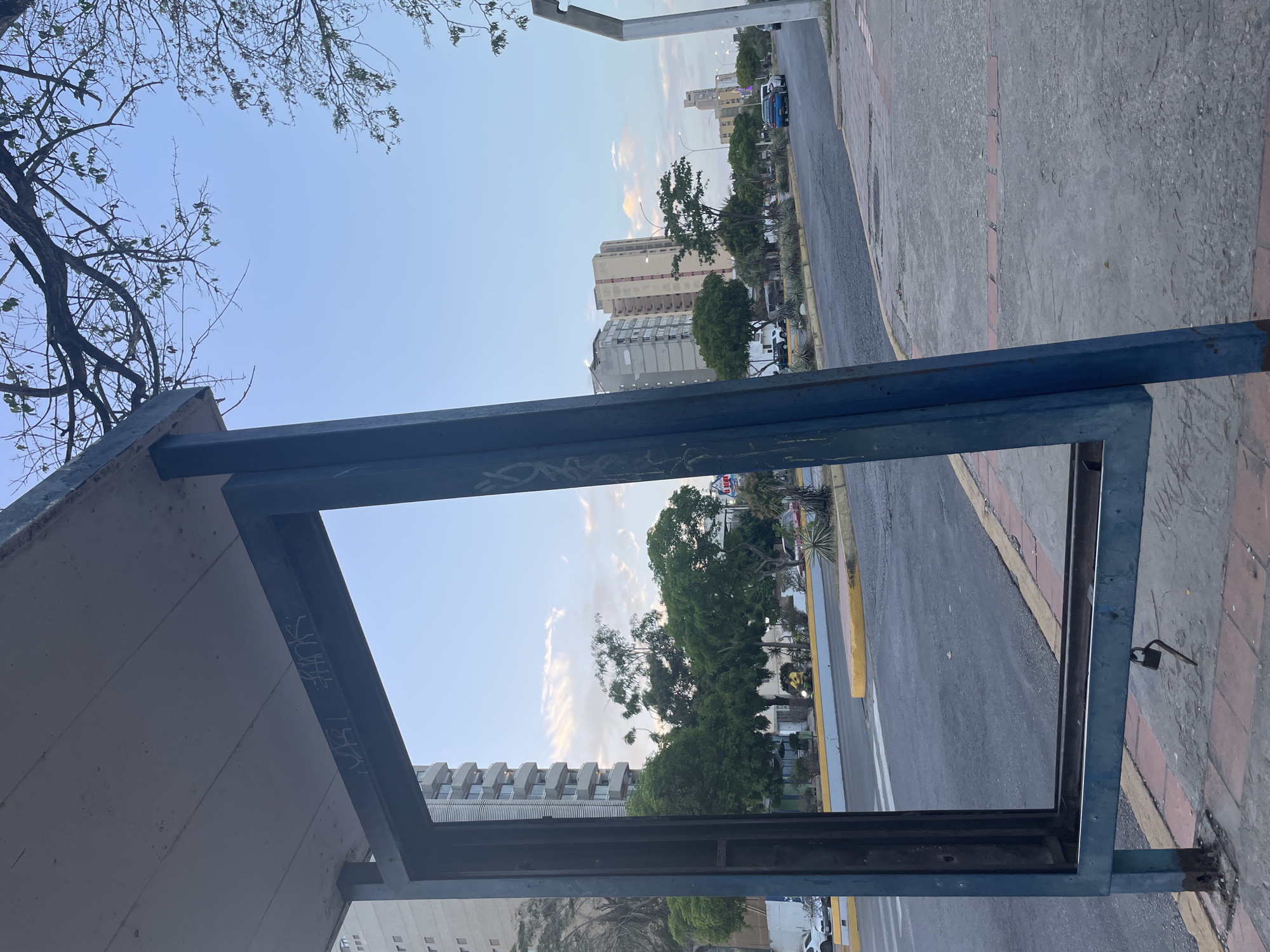 Captured from a bus stop on a Porlamar, Venezuela street, the frame shows an urban environment. Buildings of varying heights and styles line the horizon, partially obscured by lush green trees. A two-lane asphalt road runs parallel to the sidewalk, its surface marked with white lines. A yellow curb separates the sidewalk from the road. The sidewalk, paved with a combination of gray concrete and red brick, is dry and slightly weathered. Overhead, the clear blue sky is dotted with scattered clouds. The structure of the bus stop, made of blue metal, dominates the foreground, its frame marred by graffiti. A small padlock dangles from the side of the bus stop.