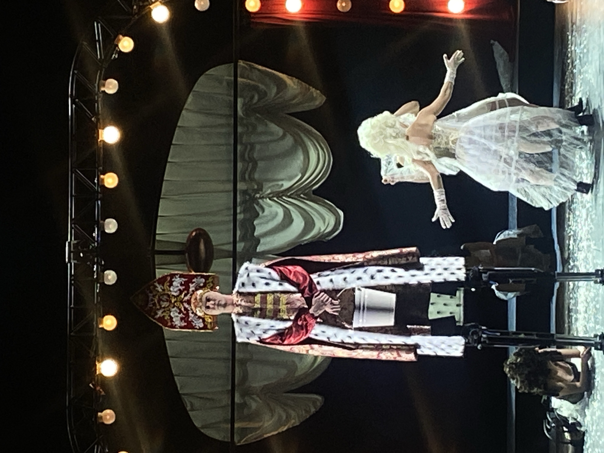 The image captures a theatrical performance, potentially in Заволжье, Russia. The focal point is a person dressed as royalty, wearing a bejeweled crown and a fur-trimmed robe, holding a silver bucket. They are positioned on a platform or stage. Above and to the side of the royal figure is a performer in a white dress and blonde wig, appearing to be in motion with arms raised. Stage lighting is prominent, featuring a metal structure with numerous round lights. A backdrop with a draped fabric design is visible behind the figures. Another person is visible kneeling on the right side of the image. The overall atmosphere suggests a dramatic or operatic scene.