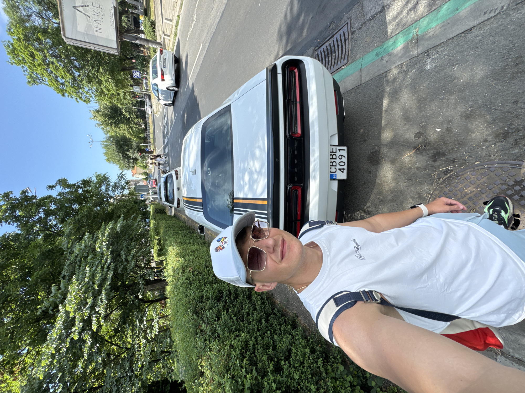 A young man wearing a white sleeveless shirt, light blue shorts, a white cap with a graphic, sunglasses, and a backpack takes a selfie outdoors. He is standing on a sidewalk next to a white Dodge Challenger with racing stripes and a Bulgarian license plate that reads "BG CBBE 4091". The car is parked on the street in front of a row of green bushes and trees. In the background, there are other cars on the road, pedestrians crossing a street, and buildings. A large billboard with the word "NAKE" is visible on the right side of the image. The scene appears to be in a city, likely Burgas, Bulgaria, given the license plate. The sky is clear and blue, indicating a sunny day.