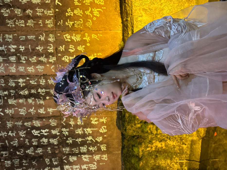 A woman in traditional Chinese attire poses in front of a stone wall with carved Chinese characters. Her elaborate headdress and gown suggest a theatrical or ceremonial context. The wall's inscriptions, arranged vertically, include phrases like "mountain city's wonder," "sun and moon eclipse," and "folk customs," suggesting themes of nature, history, and cultural heritage. The image, lit with a warm, golden glow, captures a blend of ancient tradition and contemporary presentation.