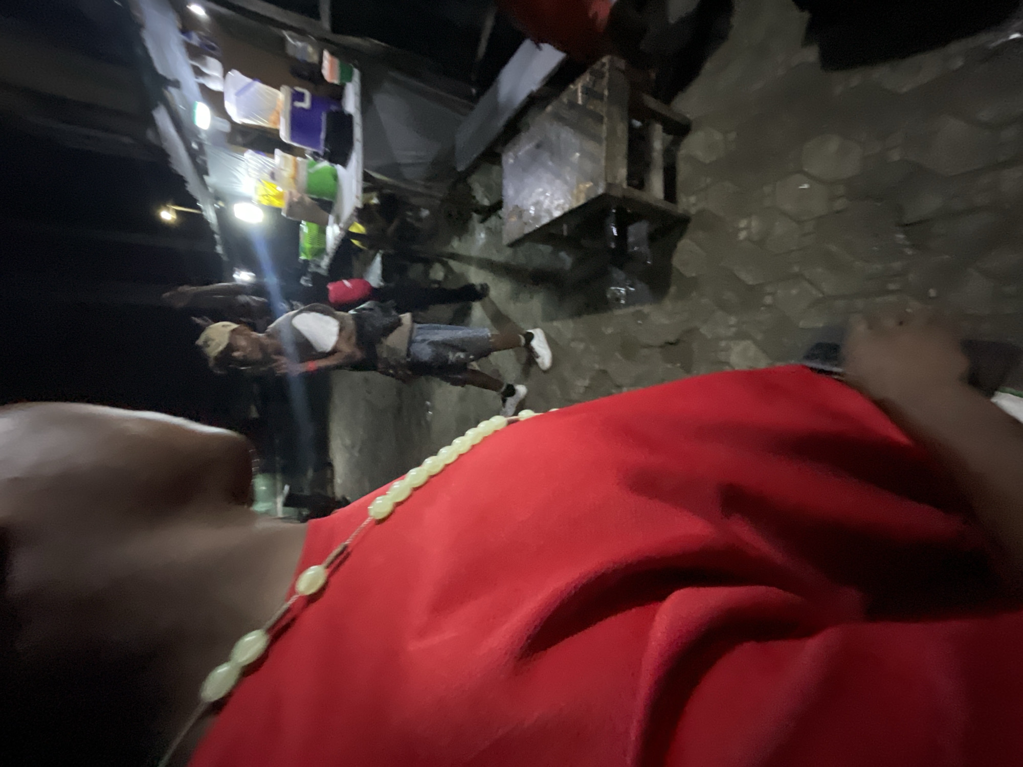 The image depicts a nighttime street scene in Ado, Nigeria, likely in a commercial or market area. In the foreground, the back of a person wearing a red shirt and a light-colored beaded necklace is visible, obscuring much of their face and body.
In the mid-ground, a second individual is standing, facing slightly away from the camera. This person is wearing a light-colored baseball cap, a grey t-shirt, and shorts. They appear to be gesturing with their left hand. Behind this person, a market stall or food vendor setup is visible. It is illuminated by artificial lighting and displays various containers, some of which appear to hold drinks or food items. A purple container and a blue cooler are prominent. A table with a metallic surface is positioned in front of the stall.
The ground is paved with hexagonal tiles, some of which appear wet or reflective, suggesting recent rain or the presence of water. The overall lighting is dim, typical of nighttime street conditions, with bright artificial lights from the stall creating localized illumination and shadows. The background is dark and indistinct, with a faint hint of street lighting further down the road. The scene suggests a casual, possibly bustling, environment at night.