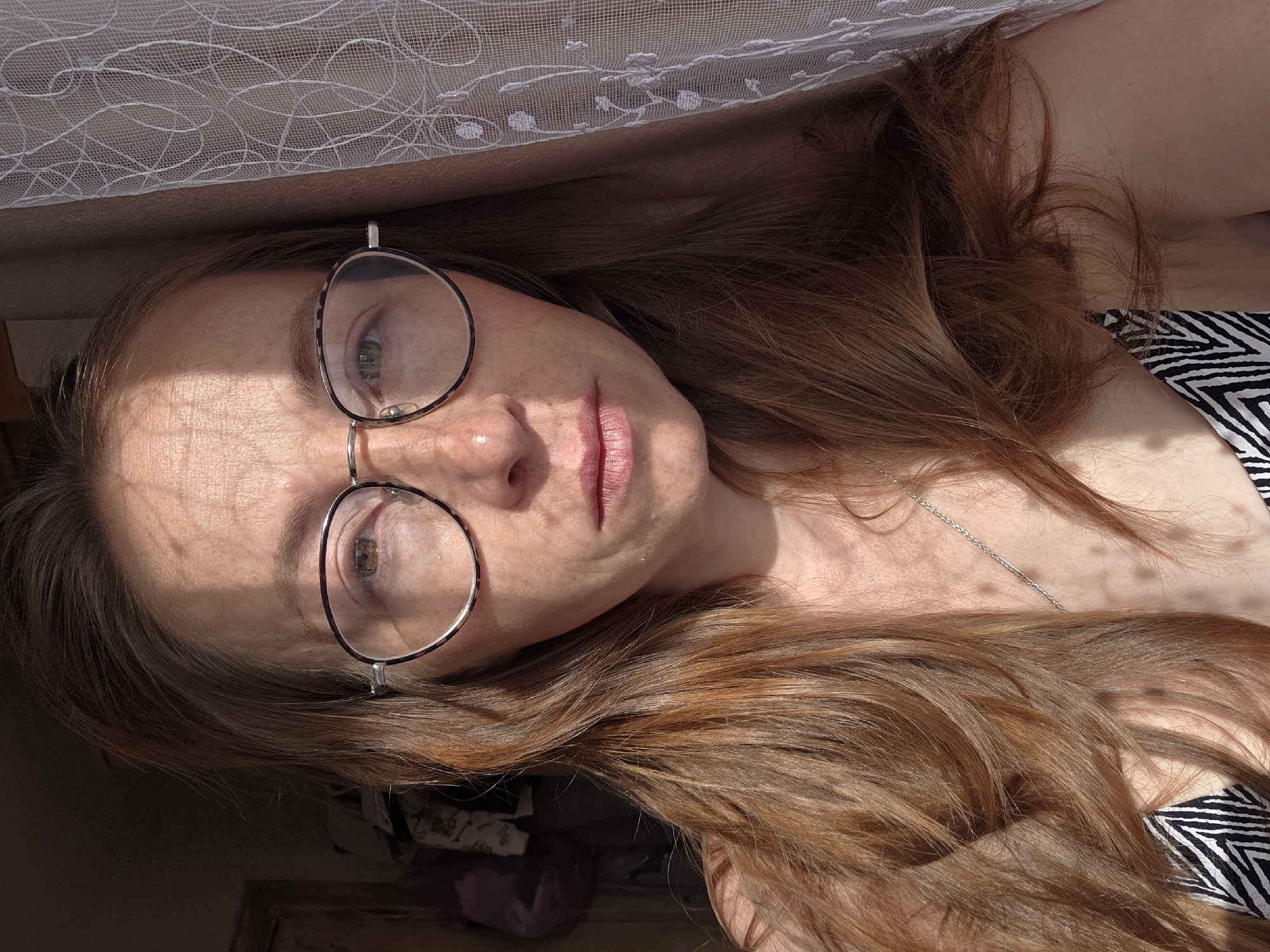 A woman with reddish-brown hair and glasses is lying down indoors near a window covered with a white lace curtain. Light is hitting her face. She is wearing a necklace and a black and white patterned garment. The location is Unknown, Unknown.
