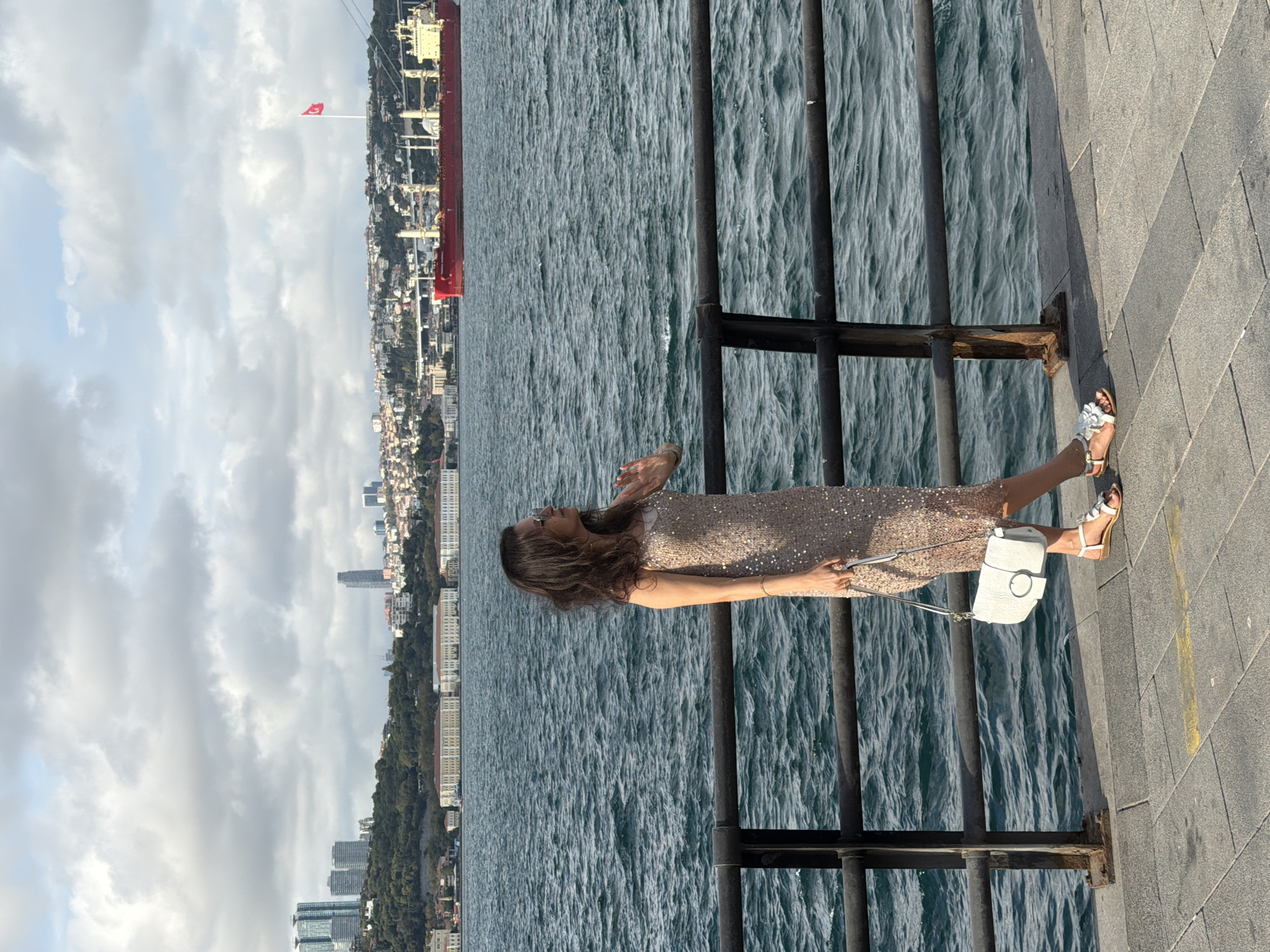 A woman stands on a paved walkway next to a dark metal railing, overlooking a choppy blue body of water. She is wearing a sequined, knee-length dress, white sandals, and carrying a white purse. The woman is looking out at the water and has her right hand raised to her face. In the background, across the water, is a city skyline with buildings of various heights and a large red cargo ship with a Turkish flag on a pole. The sky is cloudy with patches of sunlight. The scene appears to be in Üsküdar, Turkey, likely along the Bosphorus strait.