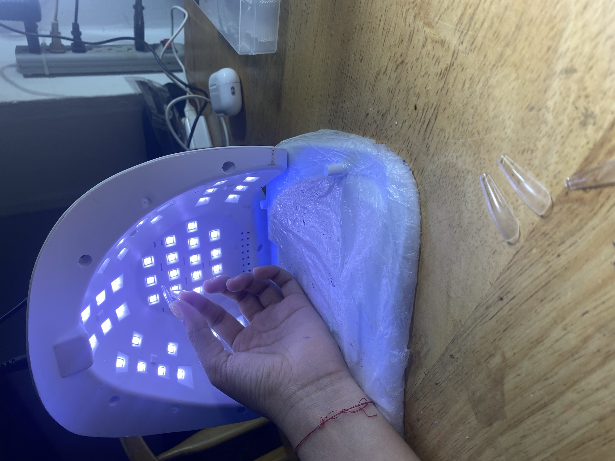 A top-down view shows a person's hand inserting a clear artificial nail into a white UV nail lamp with blue lights. The lamp is on a wooden surface. To the right of the lamp are two additional clear artificial nails. In the background, there is a power strip with plugs and a small white electrical device. The scene is indoors. The city of Cotoperíz, Venezuela is not discernable from the content of this image.