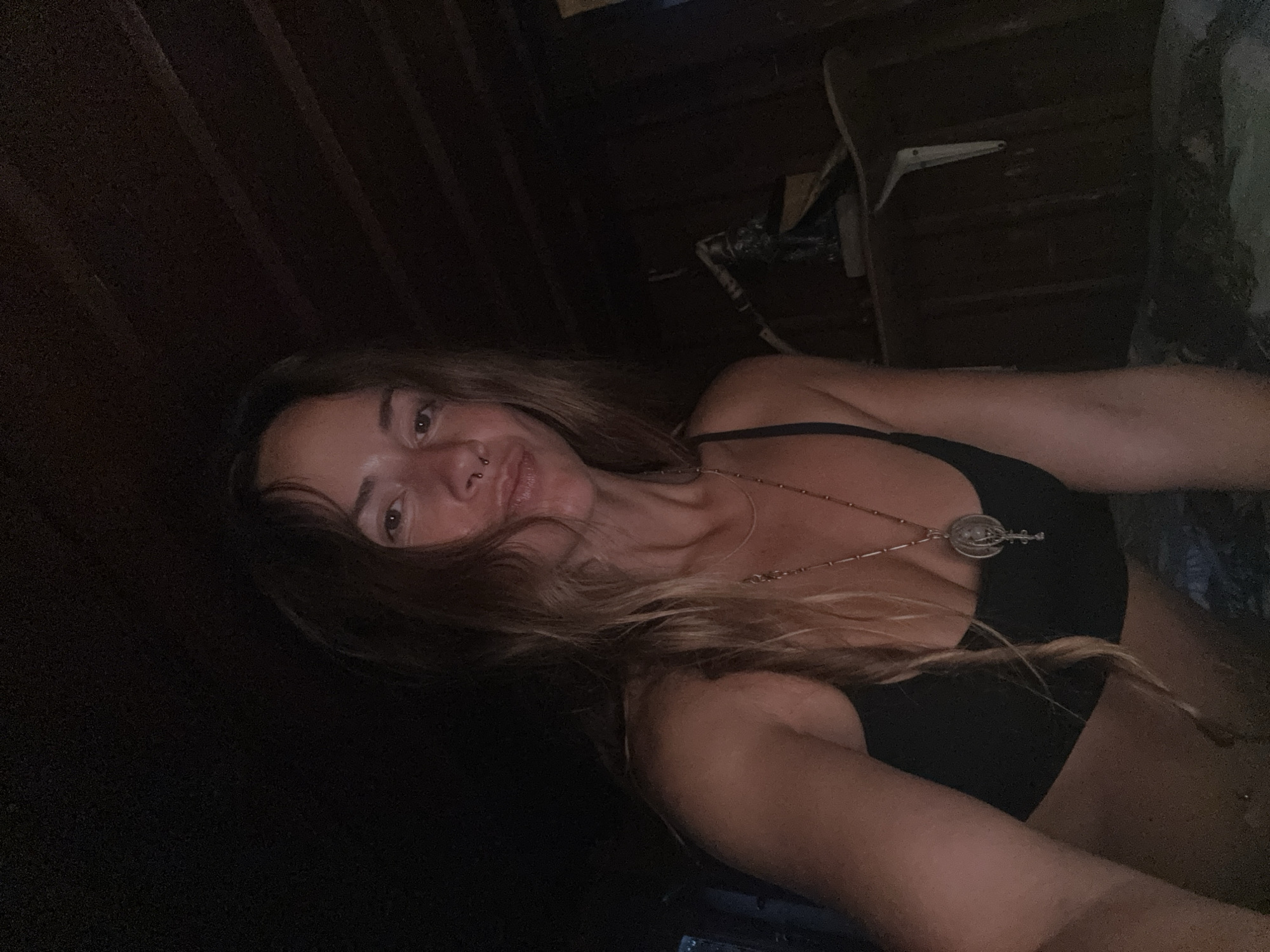 A woman with long, wavy brown hair and a nose ring is looking at the camera in a dimly lit room with wooden walls and a wooden ceiling. She is wearing a black bralette and a necklace with a circular pendant. To her right, a small statue of a person is visible on a shelf.