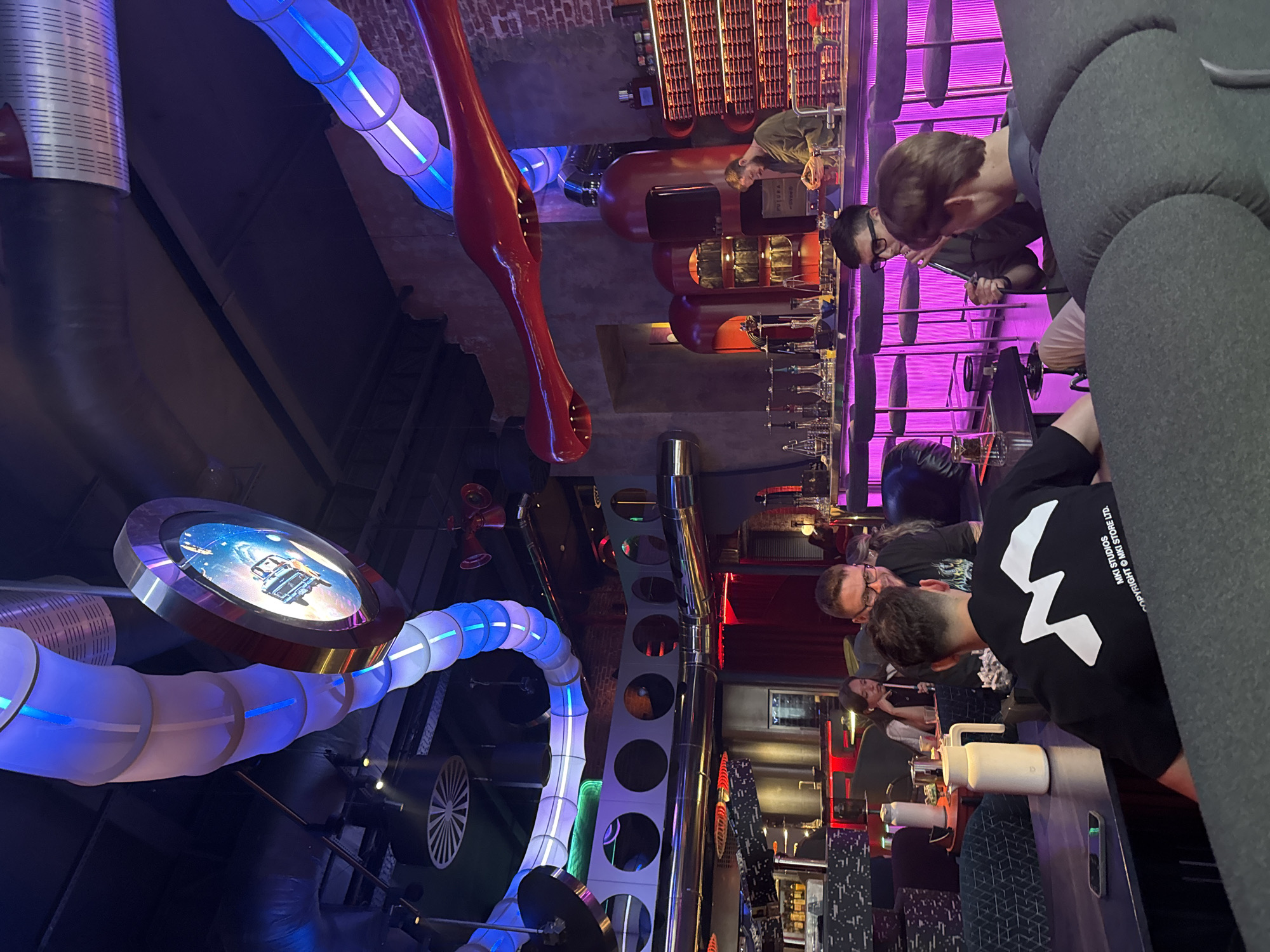 The image depicts the interior of a dimly lit establishment with a unique, futuristic decor. Suspended from the ceiling are large, segmented tubes illuminated with blue and white lights, creating a tunnel-like effect. A circular screen showing a scene from outer space is also visible overhead. The walls are a mix of exposed brick and concrete, with some sections painted a rusty red.
In the foreground, several people are seated around tables and at a bar. On the left, a person in a black t-shirt with a white logo is visible from behind, sitting at a table with a phone and a white thermos. To their right, a woman with dark hair is looking down, and a man with glasses is talking to someone behind him.
The bar area is illuminated with vibrant pink lights, highlighting rows of bar stools and the bartender, a man with a beard wearing a green shirt. He is seen working behind the bar, which is lined with shelves stocked with bottles and other items. In the background, more people are seated at tables, and the overall atmosphere suggests a modern lounge or nightclub. The presence of shisha pipes on the bar indicates it may be a hookah lounge. The style of the interior and the specific elements, such as the ventilation-like tubes and the modern furnishings, are characteristic of venues like "Fusion Fun Kitchen" in Moscow, Russia, known for its themed interior design.