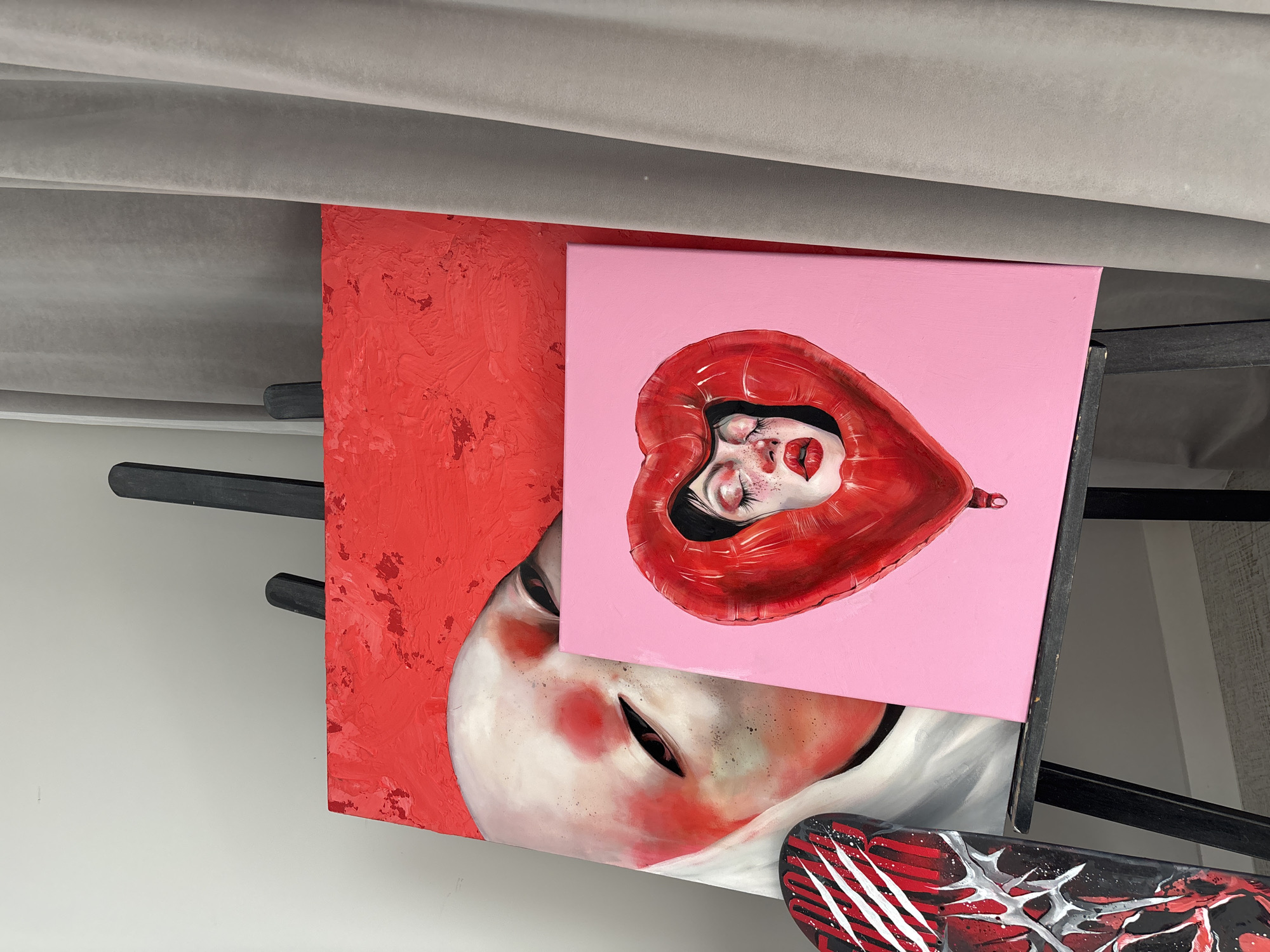 This media file captures an indoor setting, likely an art studio or display space, showcasing several artworks. The central and most prominent piece is a painting on a pink canvas depicting a person's head, with eyes closed and red lipstick, encased within a glossy, reflective red heart-shaped balloon. This pink artwork partially overlaps a larger, textured red painting to its left, which features a pale face with pronounced blush on its cheek and dark eyes.
Below the central pink painting and towards the bottom right, a segment of a black object, possibly a skateboard, is visible, adorned with dynamic red and white abstract designs. The paintings are supported by a black easel or stand, parts of which are seen behind and to the right of the canvases. The background includes a plain white wall and, at the top, a soft, folded gray material, possibly curtains or drapes. The overall impression is artistic and surreal. There is no visible text in the image.
