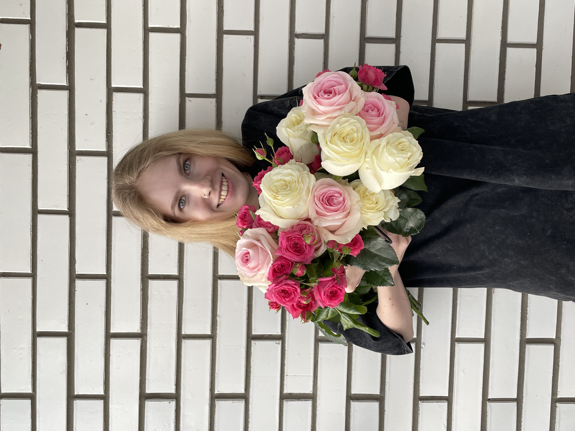 A smiling young woman with fair skin and blonde hair is holding two bouquets of flowers in front of a white brick wall. The flowers are a mix of light pink, dark pink, and cream-colored roses. She is wearing a dark-colored, textured garment. The photograph is oriented at an angle, so the brick wall appears to run vertically. The location is Izhevsk, Russia.