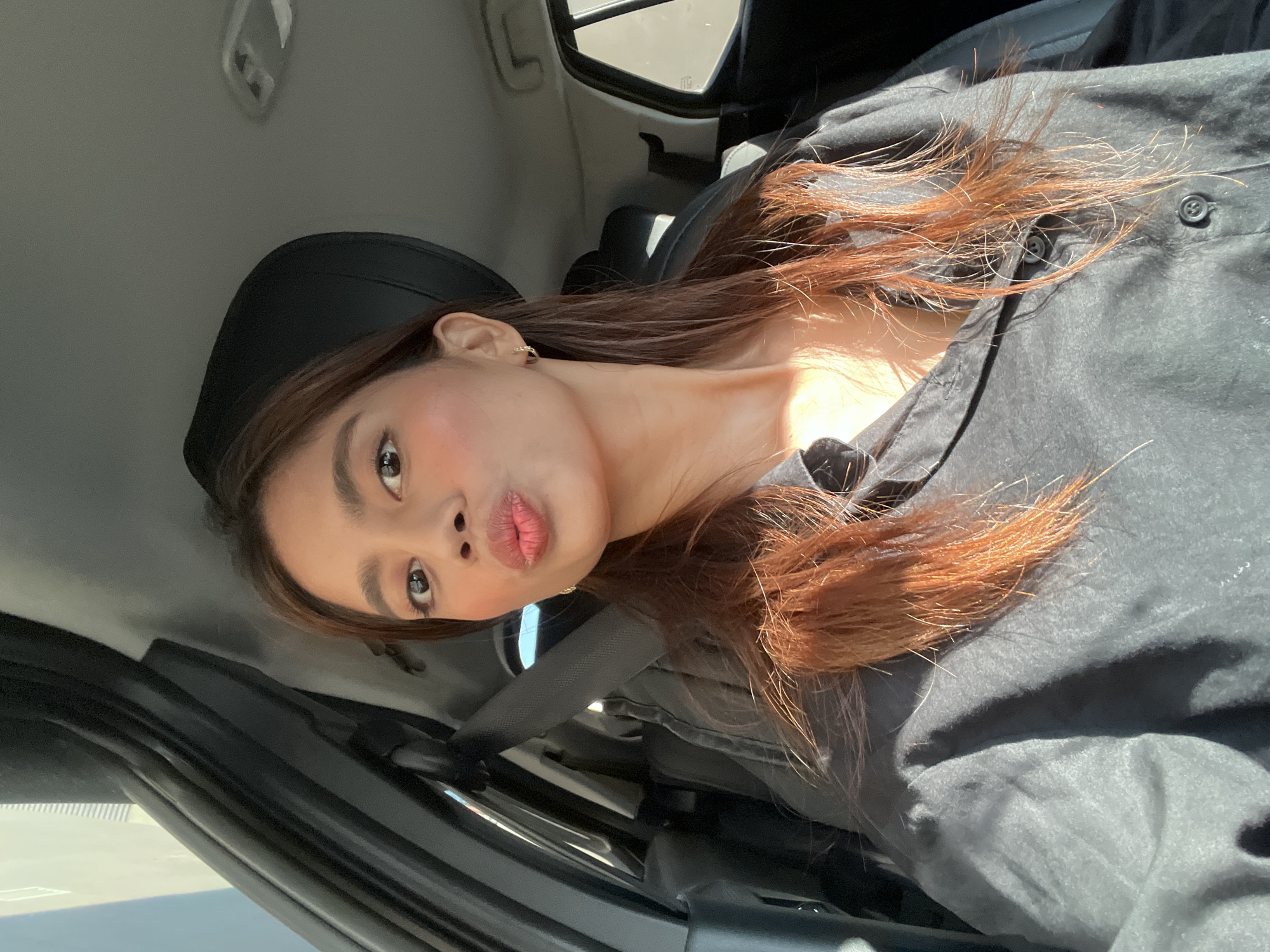 A young adult female is depicted in a selfie taken inside a car in Davao City, Philippines. She is seated in what appears to be the driver's seat, with a grey seatbelt fastened across her chest. Her dark brown hair, featuring prominent reddish-orange highlights from the sun, falls over her shoulders. She is wearing a dark, possibly black or charcoal grey, long-sleeved button-up shirt. The woman is making a pouting or "kissing" face, looking directly at the camera. Her facial features include dark eyes, defined eyebrows, and subtle blush on her cheeks. Small, gold-colored hoop earrings are visible on both ears. The car's interior shows dark upholstery on the seats and door panels, with a lighter grey headliner. An overhead interior light fixture is visible on the ceiling in the upper right portion of the frame. Strong natural sunlight enters the vehicle from the left, illuminating the subject and parts of the interior.