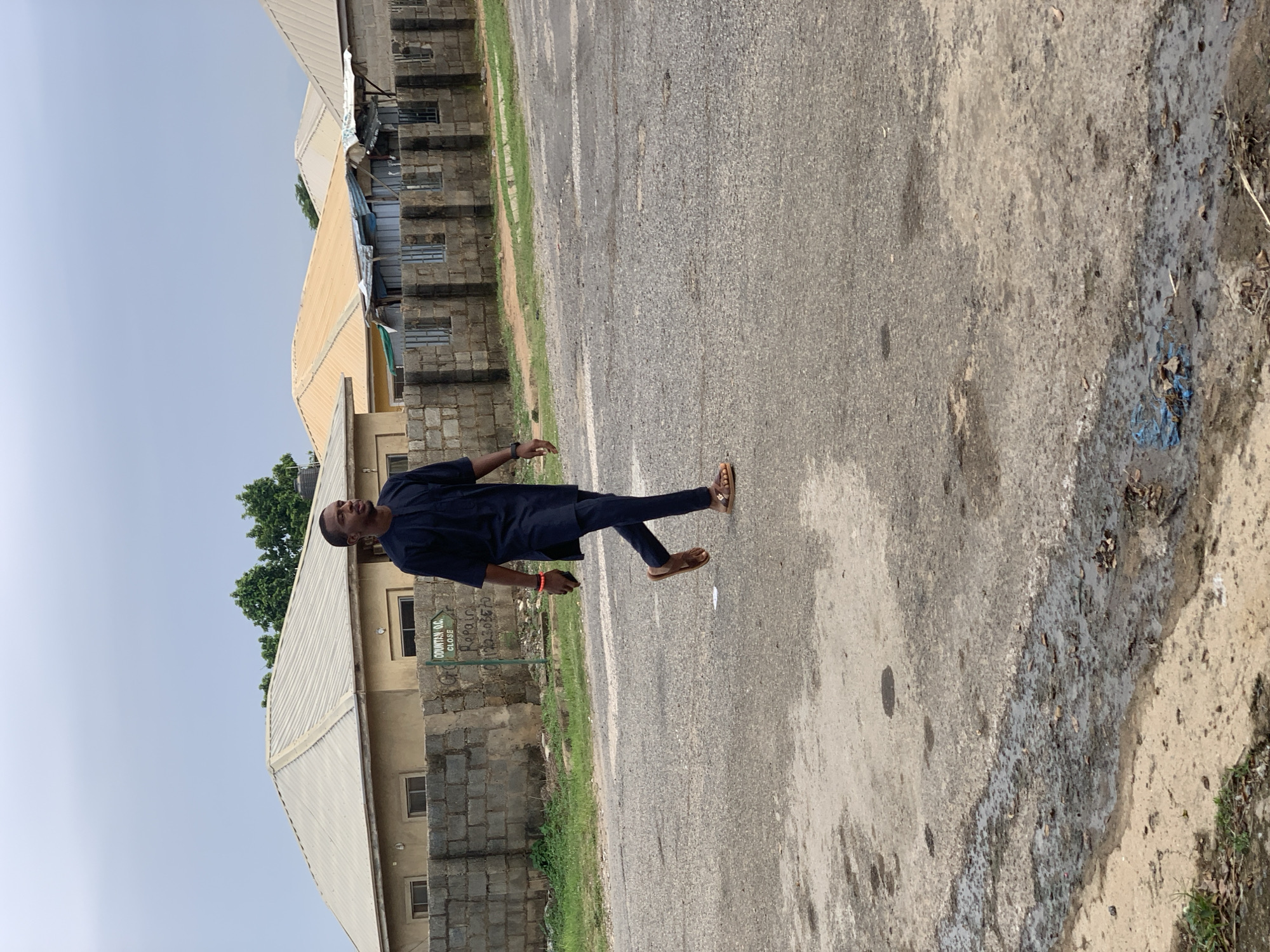 A young African man wearing a dark blue traditional outfit walks across a paved road. He is facing right, looking towards the right side of the frame. He is wearing sandals. Behind him is a low brick wall, and behind that are several single-story buildings with corrugated metal roofs. One building has a sign that reads "ODUNTAN O.C. CLOSE" and "Repair". The sky is overcast. The scene appears to be in a residential area in Abuja, Nigeria.