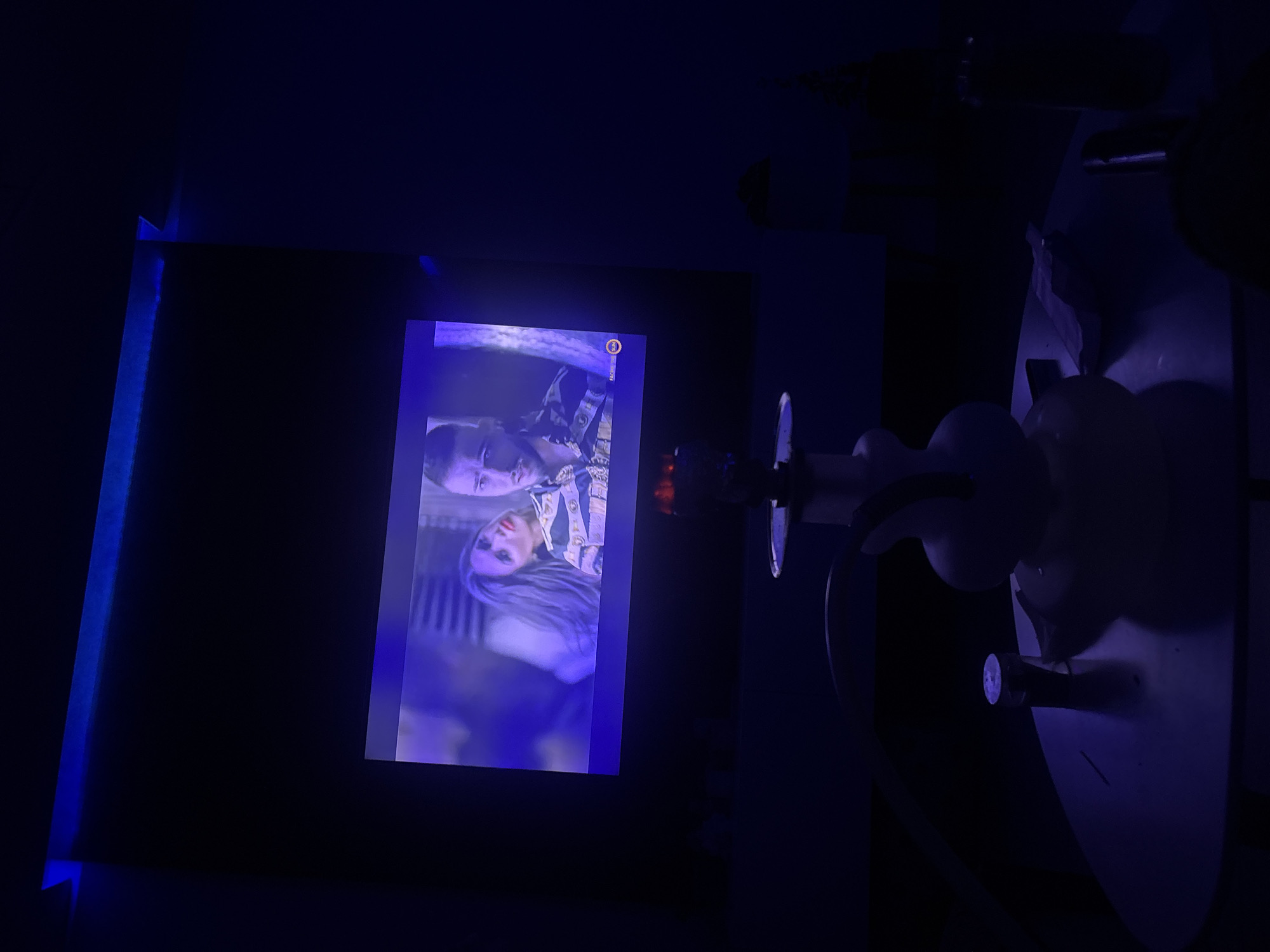 A hookah sits on a table in front of a large screen displaying a movie. The room is dimly lit with blue LED strip lights illuminating the top edge of the screen. The screen shows a close-up of a man and a woman, with the woman appearing to be leaning on the man's shoulder. The man is looking directly at the camera with a serious expression, and the woman has her eyes closed. A water pipe, commonly known as a hookah, is prominently positioned in the foreground, centered on a circular table. The hookah has a white base and a clear stem. A small cylindrical object, possibly a lighter or a measuring tool, is placed next to the hookah on the table. The overall ambiance suggests a relaxed and intimate setting, likely for entertainment and social gathering.