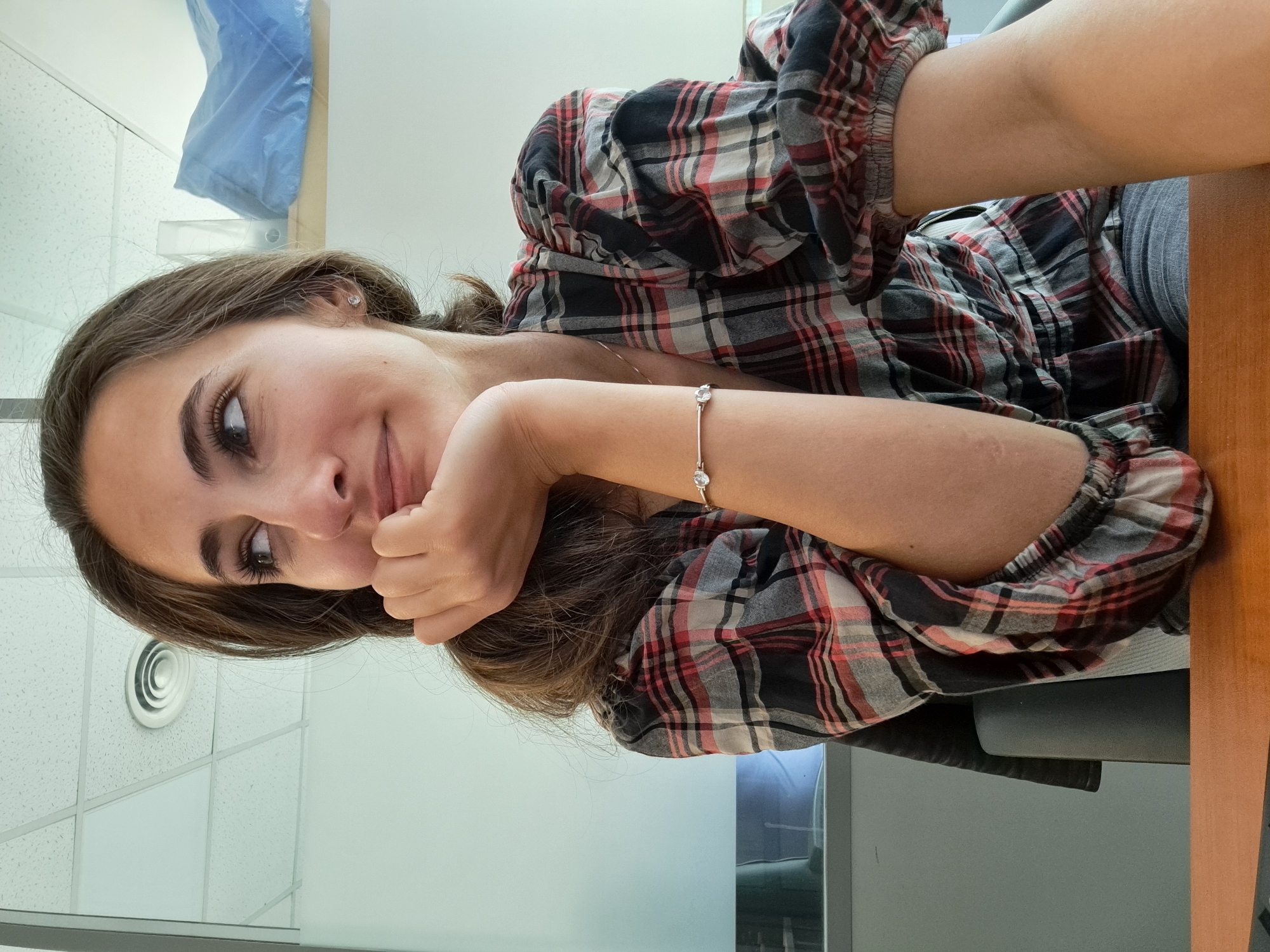 A young woman is posing for a selfie indoors. She is fair-skinned with brown hair and wearing a red, black, and white plaid shirt. She rests her head on her hand, and a gold bracelet is visible on her wrist. The background shows a white, drop-ceiling panel and a portion of a wooden desk. The setting cannot be conclusively determined as Sofia, Bulgaria from the provided image.