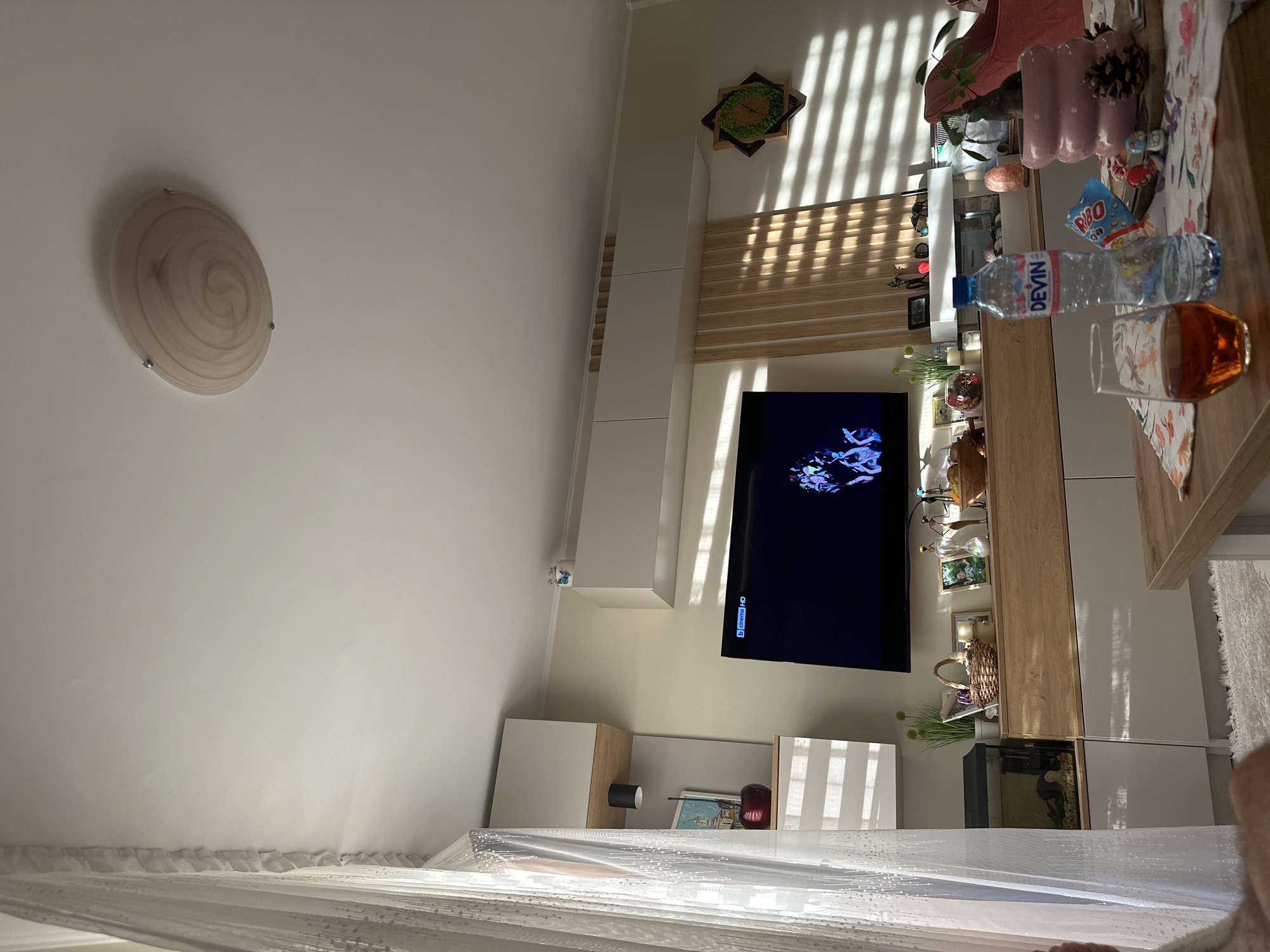 A slightly rotated interior shot of a room, likely in Plovdiv, Bulgaria, shows a living space with a modern aesthetic. Dominating the wall is a flat-screen television displaying an animated scene, mounted above a modular entertainment unit featuring a blend of white and light wood finishes. The unit incorporates open shelves, cabinets, and an aquarium. Various decorative items and personal effects, such as vases, picture frames, and bottles, are arranged on the shelves and surfaces. To the left of the TV wall is an off-white wall with a decorative ceiling light fixture. Natural light streams through a window covered by a sheer white curtain, casting shadows across the room. In the foreground, a portion of a table is visible, with household items like water bottles, a glass, and decorative objects placed on it.