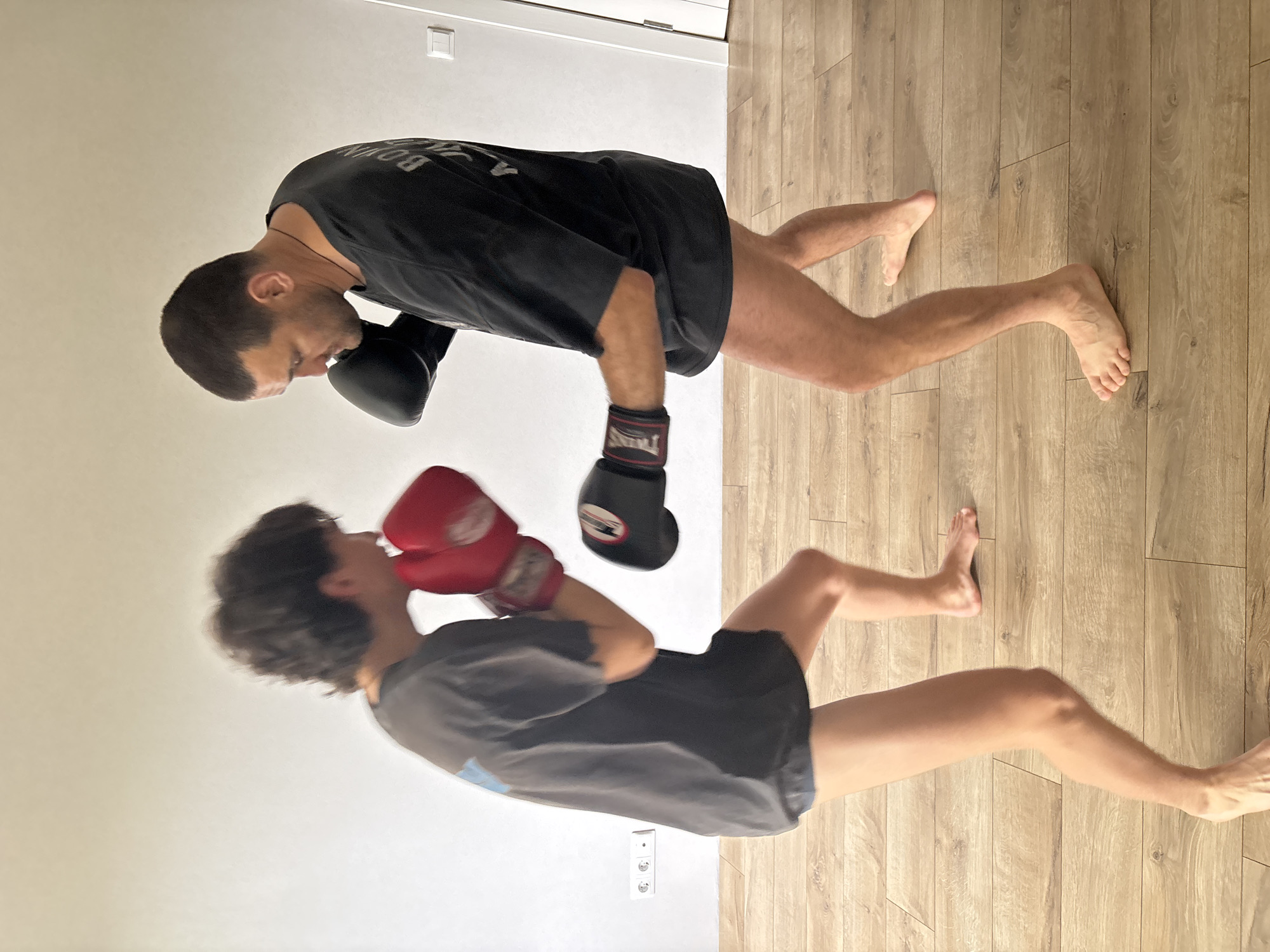 Two individuals are depicted indoors, engaged in a boxing or martial arts training session. The image is rotated 90 degrees counter-clockwise, showing a white wall on the left and a light-colored wooden floor on the right.
One individual, with short dark hair, wears a black t-shirt with the word "BOXING" visible on the back, black shorts, and black boxing gloves. The brand "TWINS" is visible on the wrist of one black glove. This person is captured in a stance with their head slightly lowered, facing the other individual.
The second individual, with curly dark hair, wears a grey t-shirt, black shorts, and red boxing gloves, also bearing the "TWINS" brand logo. This person is also in a dynamic stance, with arms raised, and appears to be in motion, as indicated by slight blurring. Both individuals are barefoot.
The scene suggests an active training moment, possibly sparring, indoors. The lighting is bright and even, indicating daytime. Visible on the white wall are a white light switch and a European-style electrical outlet. The location context is Chaikovsky, Russia, which aligns with the style of the electrical outlet. Emotions are not clearly discernible, but concentration and physical activity are implied by their stances.