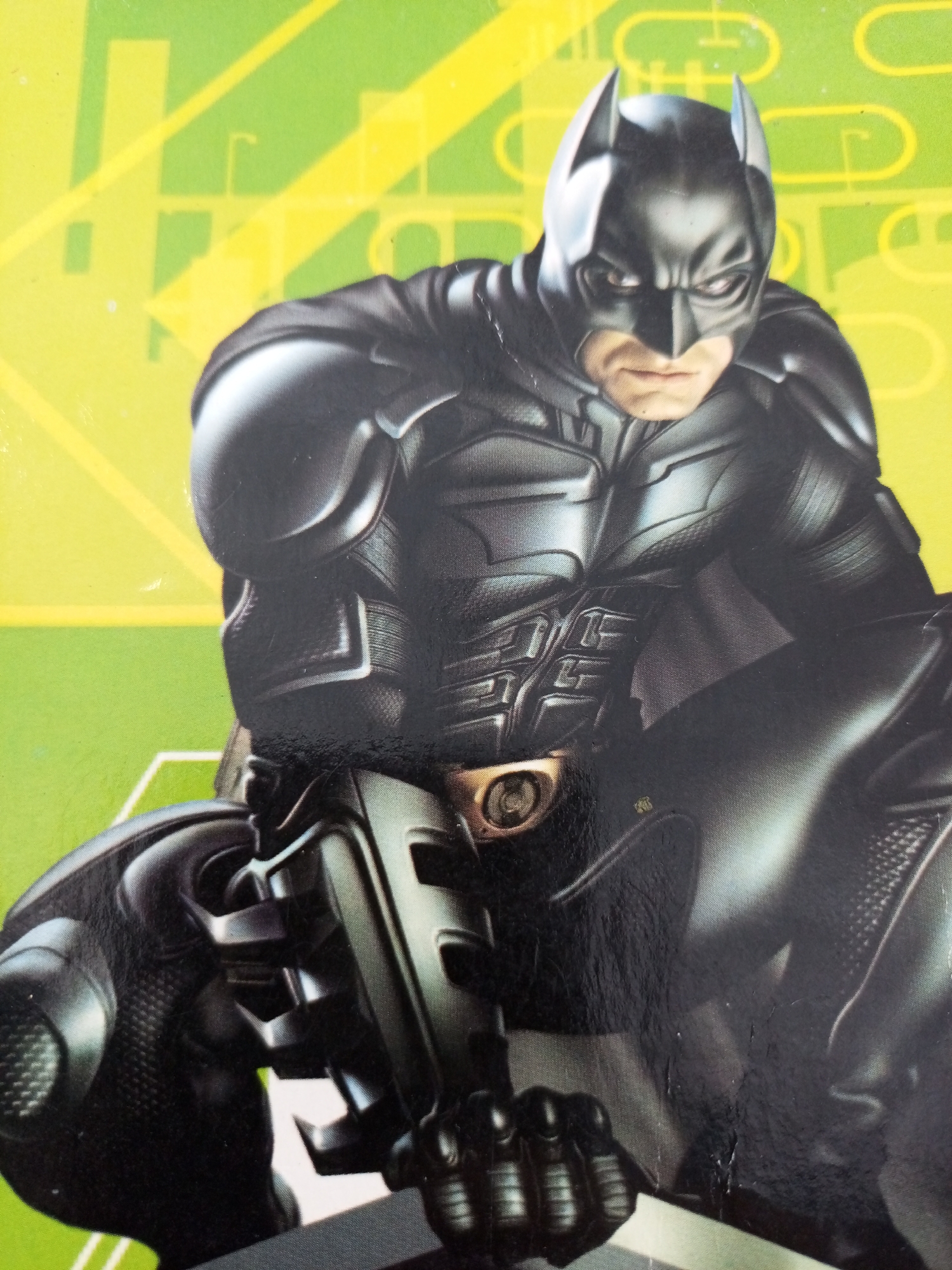 The image shows a close-up, low-angle shot of a figure in a black Batman costume, which appears to be a statue or action figure. The figure is positioned in a crouched stance, facing slightly to the right. The costume features intricate detailing, with segmented armor plating on the chest, shoulders, and legs. A prominent belt with a circular buckle is visible at the waist. The background is a vibrant green and yellow abstract pattern, possibly representing a city skyline or geometric design. There are no discernible elements in the image that would allow for the identification of the location in Lhokseumawe, Indonesia.