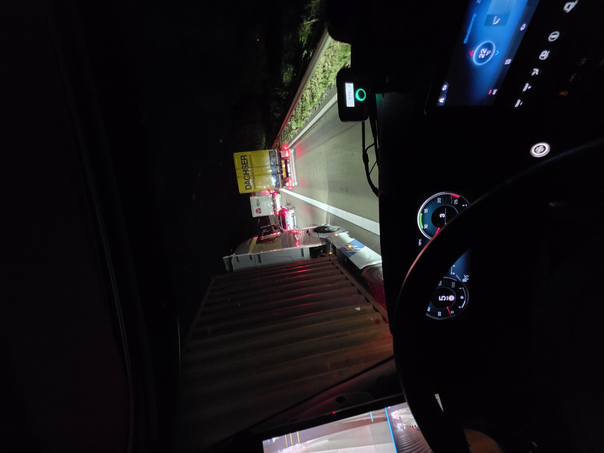 The image captures a view from the driver's seat of a truck at night, looking forward and slightly to the right, showing a traffic jam on a highway. In the foreground, the steering wheel and dashboard are visible, with illuminated gauges displaying speed and gear (indicated as "5"). To the right, a digital screen shows "Klimatisierung" (climate control) with a temperature of 22°C. The truck is stopped or moving very slowly behind another large truck with a yellow "DACHSER Intelligent Logistics" trailer. Further ahead, other vehicles, including a white DPD truck and cars, are visible with their brake lights illuminated. The highway has a concrete barrier on the right side, with vegetation beyond it. The scene appears to be on the Autobahn A3 near Rodgau, Germany, based on the context of the vehicle and the general appearance of German highways at night. There are no people clearly visible outside of the assumed presence of the driver.