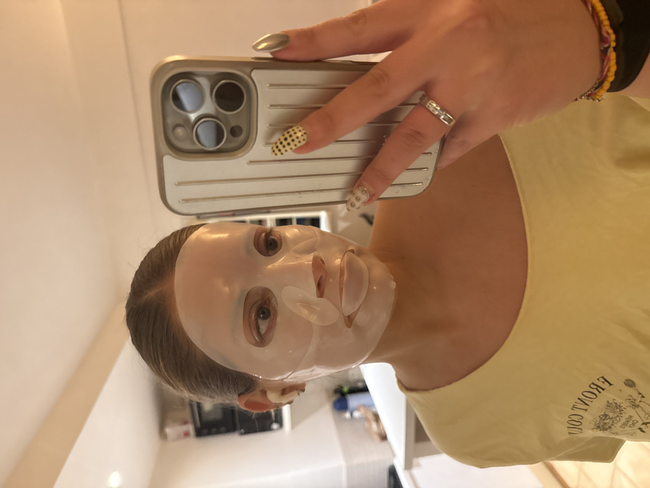 A person wearing a face mask takes a selfie with a silver iPhone. They are wearing a light yellow shirt and a ring on their left hand. Their fingernails are manicured, with one fingernail painted silver and another painted with black polka dots. A colorful bracelet is visible on their wrist. The background appears to be a kitchen area.