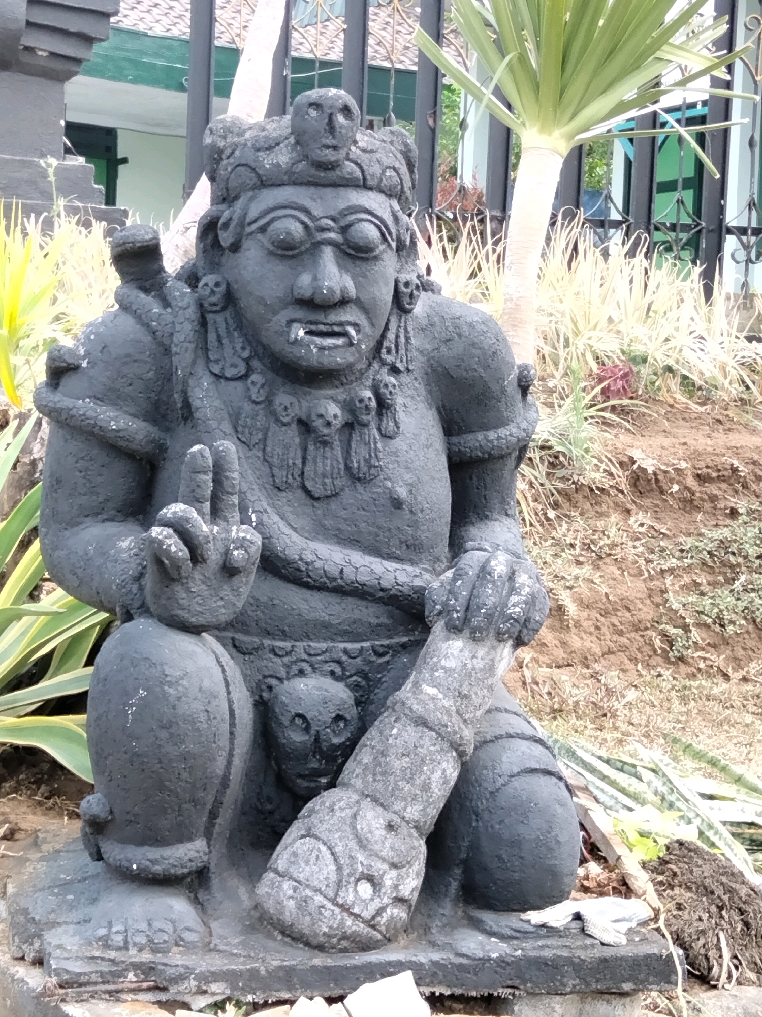 This is a waist-up photograph of a dark grey, weathered stone statue depicting a seated figure. The statue is in a crouched position, with its left leg bent and its right leg tucked beneath it. The figure has a fearsome expression, with wide eyes, bared teeth, and a prominent forehead. It wears a headdress adorned with skull-like carvings and appears to have snakes coiled around its neck and shoulders. The figure's right hand is raised, showing two fingers extended. A carved phallic object rests on its lap, with a smaller skull-like carving positioned below it. The statue is situated outdoors on a stone base, with sparse vegetation and a dirt embankment in the background. To the left of the statue, a portion of a dark grey building with a tiled roof is visible. The overall scene suggests an outdoor decorative element in a garden or temple setting. The statue is likely located in Batu, Indonesia, a city known for its numerous stone sculptures and attractions.