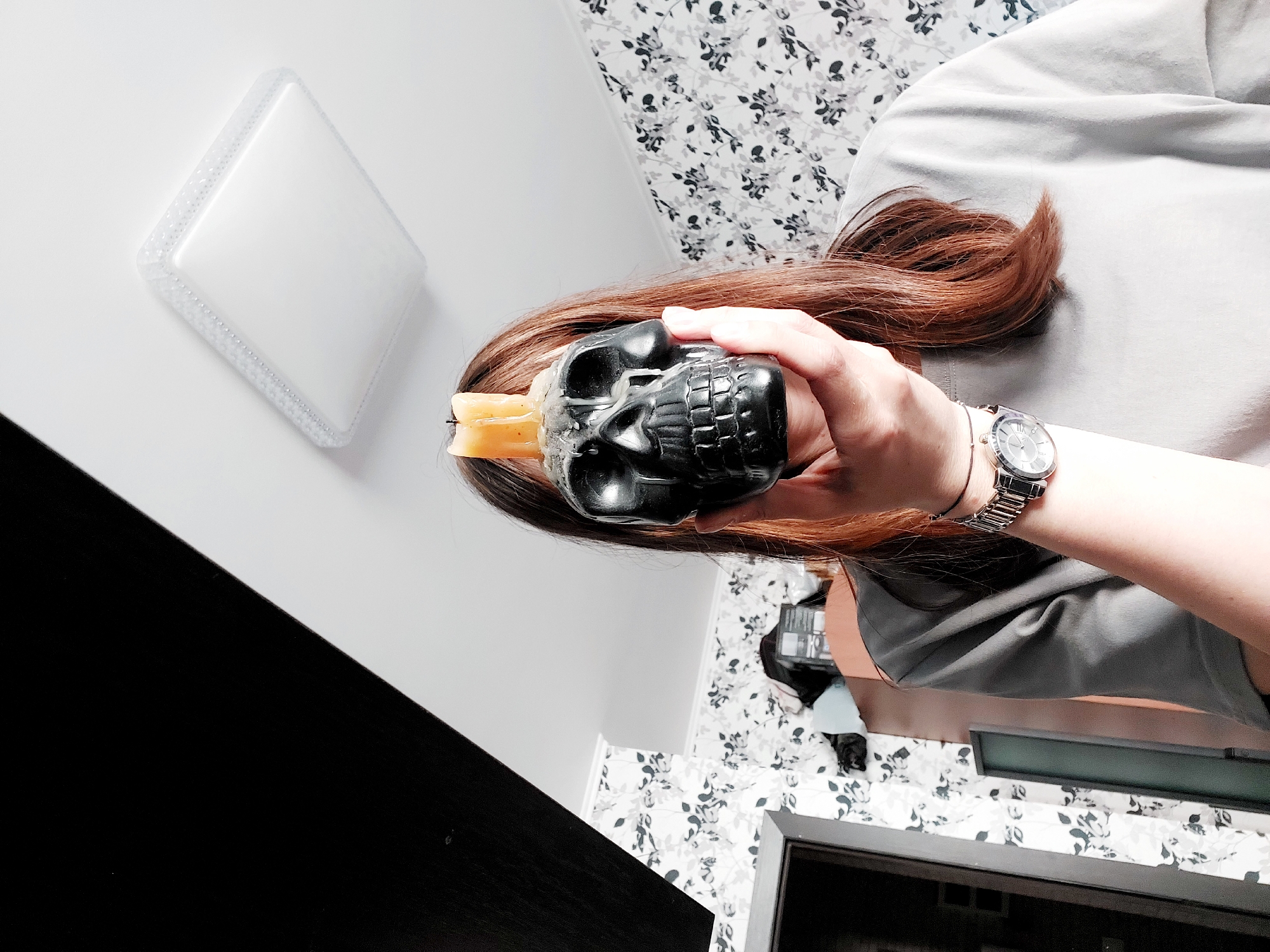 A person wearing a watch is holding a skull-shaped candle holder in their left hand. The candle holder is black and has two yellow candles inserted into it. The person has long, brown hair. A white square light fixture is visible on a white surface. The background shows a white wall with a black floral pattern. A black surface and an object with a floral pattern are also visible. The location of the image is unknown.