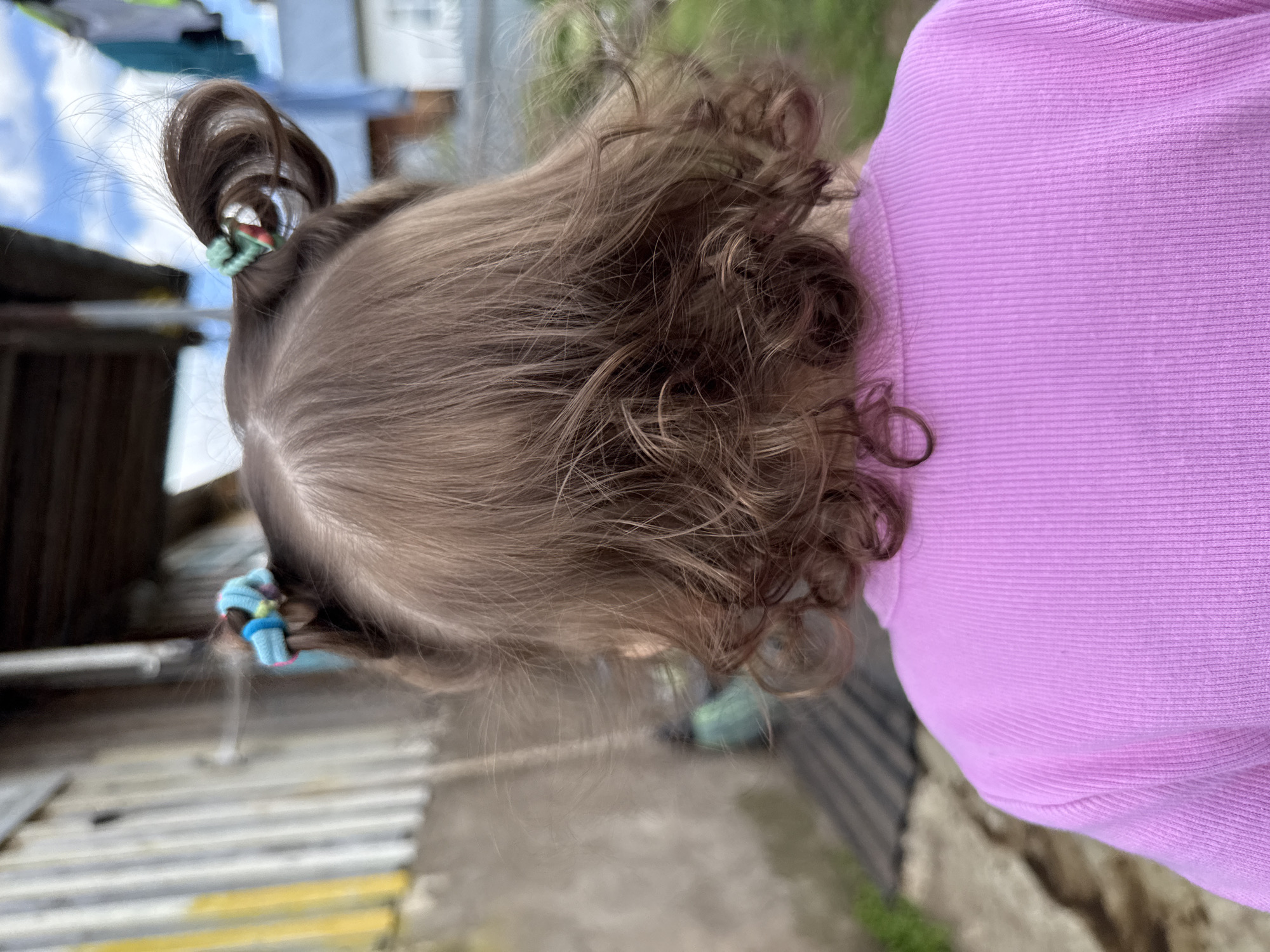 A close-up, slightly blurred, outdoor shot shows the back of a young child's head and shoulder, with the focus primarily on the child's hairstyle. The hair is light brown, styled in two high pigtails secured with colorful hair ties. The hair at the nape of the neck is curly. The child is wearing a pink sweater. The background shows a blurred, indistinct urban environment with hints of buildings, possibly a fence, and sky. The photo was taken outdoors in Уральское, Russia.