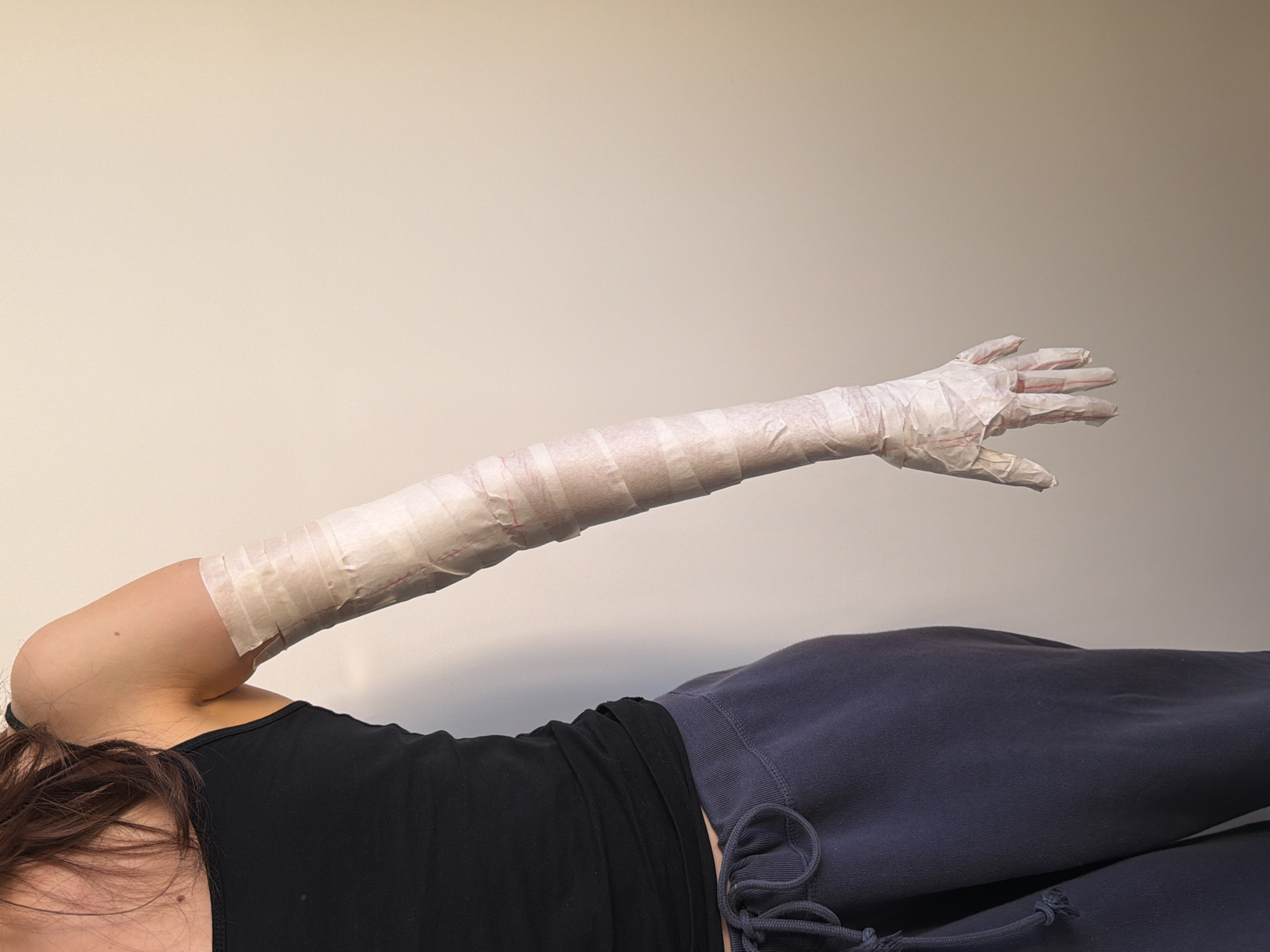 A person wearing a black tank top and navy blue sweatpants is lying down with their left arm extended upward. The arm is wrapped in strips of white tape from the shoulder down to the hand, effectively creating a cast. The person's head is partially visible, with brown hair partially obscuring the face. The background is a plain, light beige wall. The location is not identifiable as Milan, Italy, from the provided image.