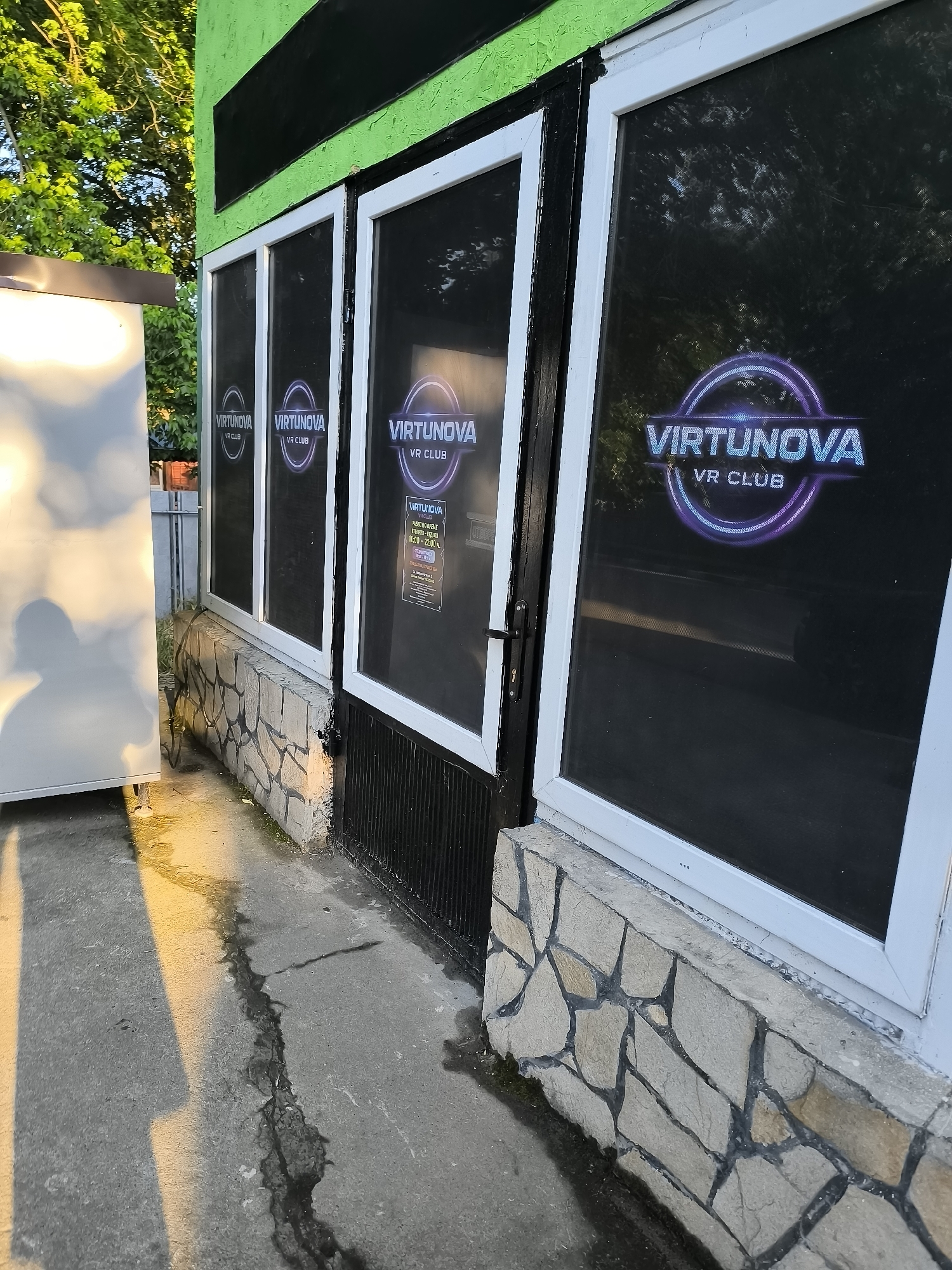 The image captures the exterior facade of a business called "VIRTUNOVA VR CLUB" in Trastenik, Bulgaria. The building features a vibrant green upper wall, beneath which are four large window panels, possibly part of a storefront or entrance. The lower portion of the facade is adorned with a decorative stone-patterned base.
The windows display prominent circular logos with the text "VIRTUNOVA VR CLUB" in a stylized, glowing font, suggesting a virtual reality entertainment venue. On one of the central window panels, a sign provides the operating hours: "РАБОТНО ВРЕМЕ" (Working Hours), "ВСЕКИ ДЕН" (Every Day), "10:00 - 22:00ч." (10:00 AM - 10:00 PM).
To the left of the building, a large, light-colored object, possibly a utility box or a temporary structure, is partially visible. Its surface reflects the warm, golden light of the sun, indicating it is late afternoon or early evening. The ground in front of the building is concrete, with a noticeable crack running down its length, and some dampness is visible near the base of the wall. Lush green foliage from trees or bushes can be seen in the background, partially reflecting in the dark window panes. The overall scene suggests a quiet moment at the entrance of an entertainment establishment as daylight begins to wane.