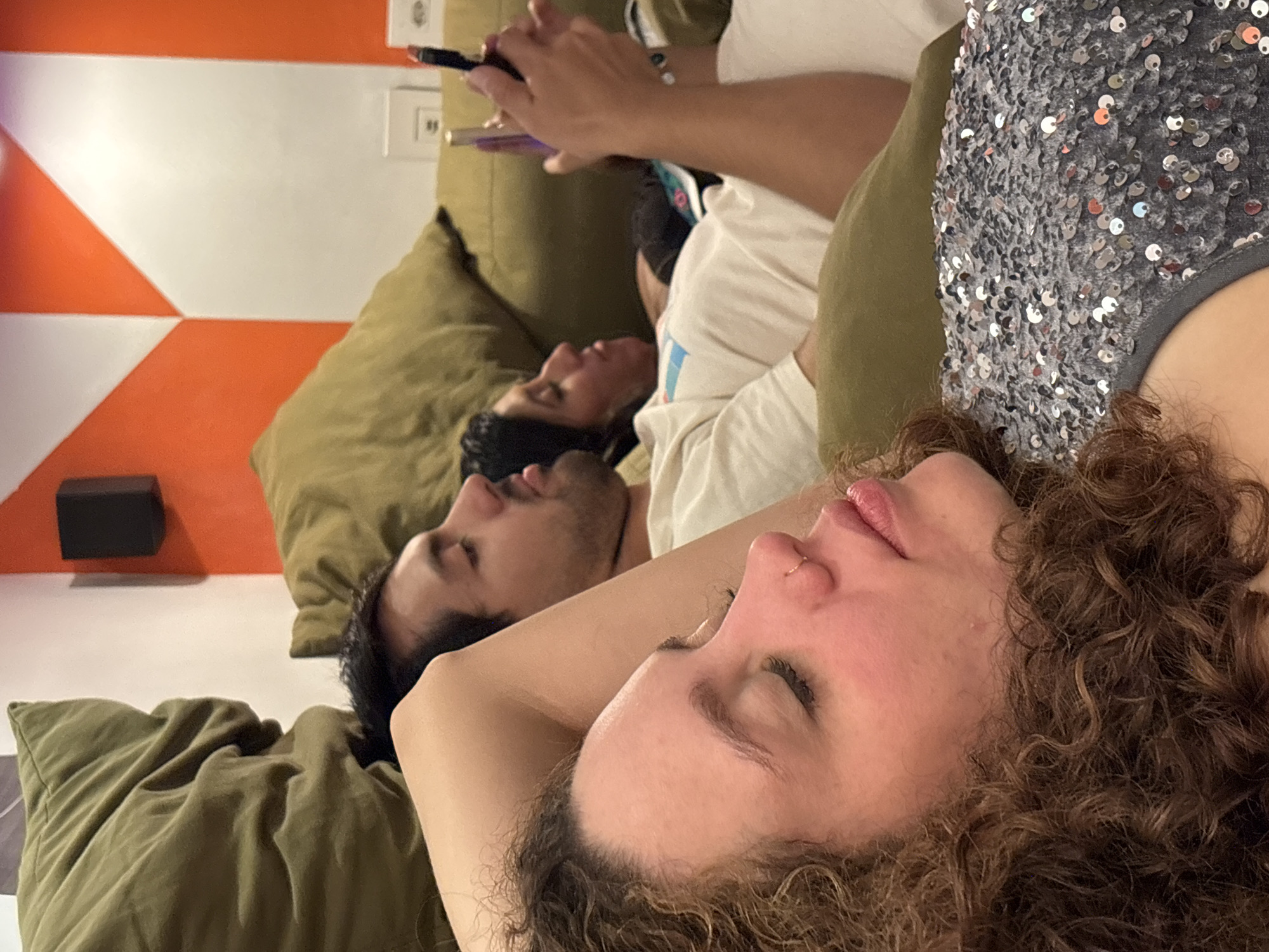 This close-up indoor shot captures three individuals relaxing, likely on a sofa or bed, from a low-angle perspective looking upwards. The setting features olive-green cushions.
In the foreground, a person with curly brown hair and fair skin is depicted with closed eyes, lips slightly parted, and a subtle nose ring, indicating rest or sleep. They are wearing a grey top embellished with silver sequins.
Behind this person, a second individual with short dark hair and a beard is also lying on their back, eyes closed, seemingly in a similar state of repose.
Further in the background, a third person, wearing a white t-shirt, is actively engaged with a smartphone, held in their hands. A light-colored bracelet or watch is visible on their wrist.
The background wall features a distinctive geometric pattern of orange and white triangles and diagonal lines. Two white electrical outlets are visible, as is a small, black rectangular object mounted on the wall. The overall atmosphere suggests a moment of leisure and relaxation. While the provided context suggests São Paulo, Brazil, there are no specific visual cues within the image itself to confirm this location. No text is visible in the image.