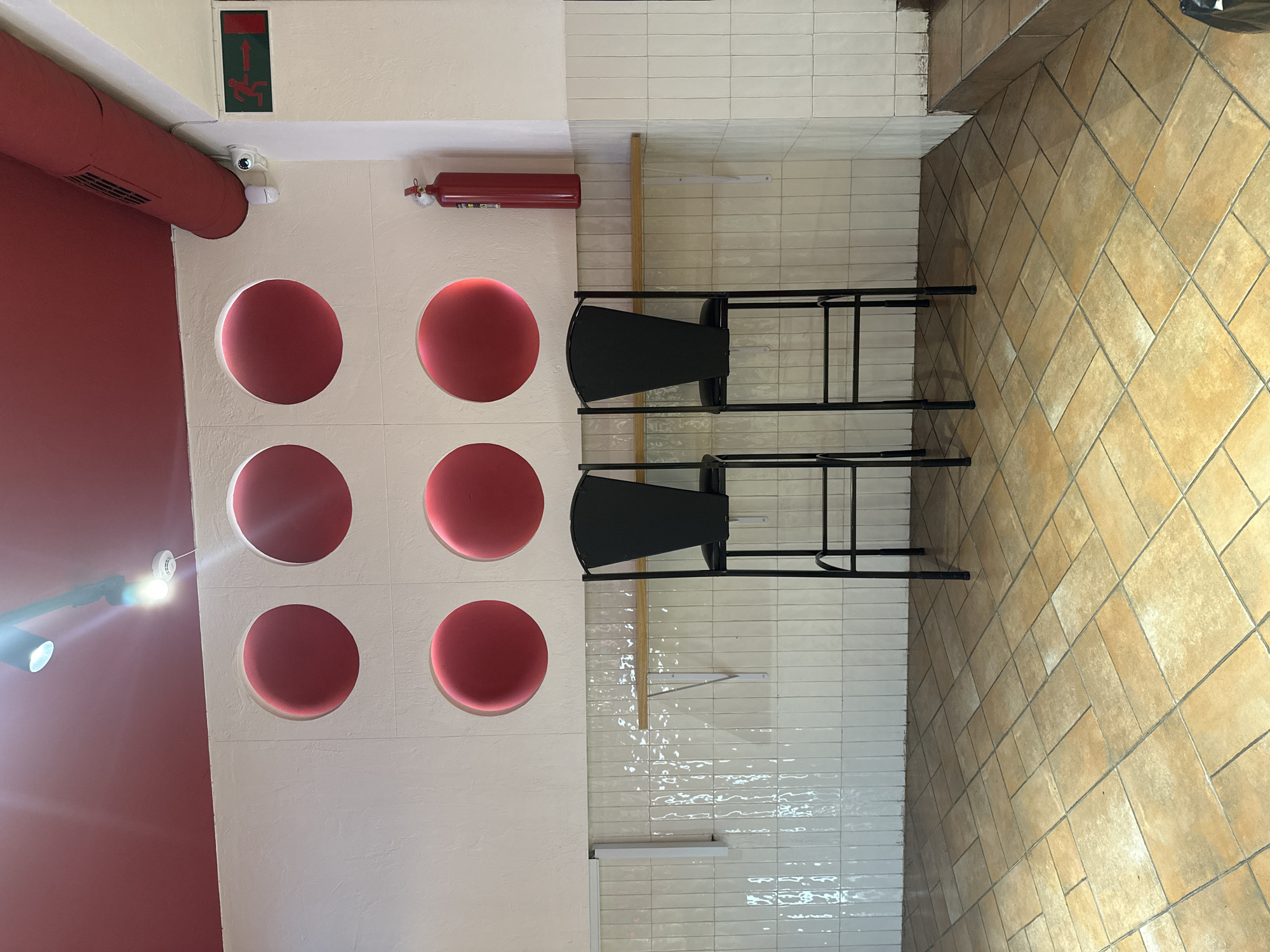 A vertically oriented, eye-level shot depicts an interior scene. On the left is a red wall and a white wall with circular cut-outs. A red fire extinguisher is mounted on the white wall, along with an exit sign. Two black bar stools are positioned near a wall tiled with narrow white tiles. To the right, the floor is tiled in a diamond pattern with light brown tiles. The location may be Yerevan, Armenia. There are no people in the shot.