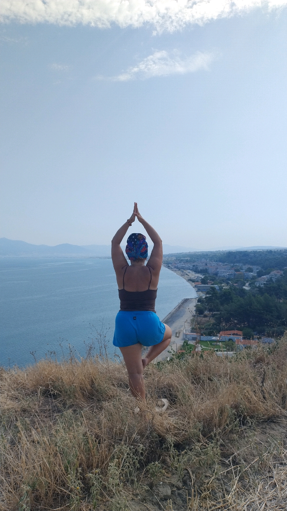 A woman is performing a yoga pose, specifically the Tree Pose, on a grassy, dry hillside overlooking the Aegean Sea in Agia Triada, Greece. She is facing away from the camera, her hands clasped together above her head, and her right leg is bent with her foot placed on her left thigh. She is wearing a dark brown tank top, bright blue shorts, and a patterned headscarf. The sea stretches out to the horizon, with a coastal town visible along the shoreline. The sky is a clear blue with a few wispy clouds. The foreground is dominated by dry, straw-like grass and some scattered rocks.