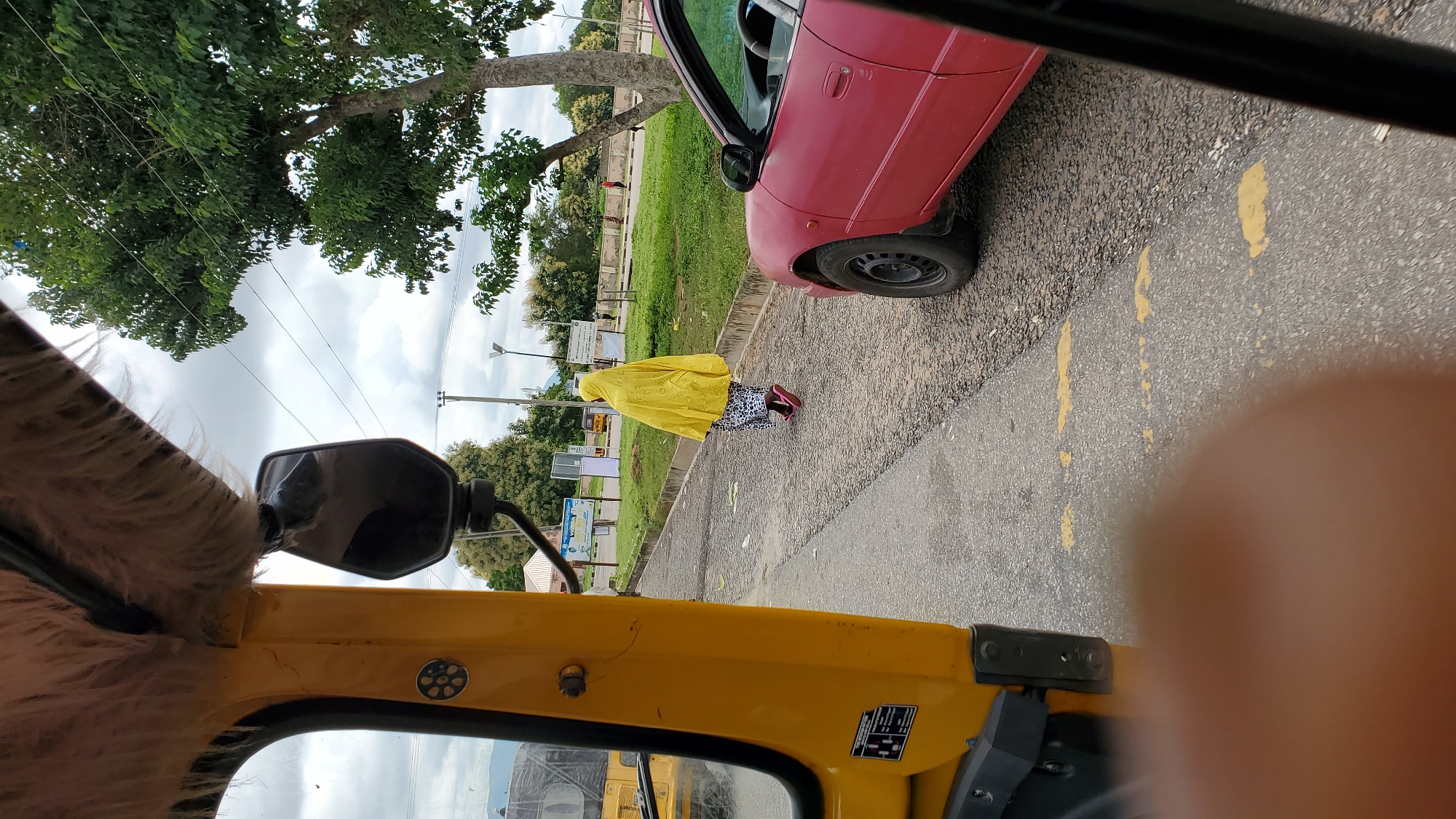 The image captures a street scene in Jalingo, Nigeria. In the foreground, a yellow vehicle, likely a tricycle taxi, is partially visible, its fuzzy interior lining and side mirror indicating the photographer's viewpoint. The road is paved with asphalt and marked with yellow lines. A person, wearing a bright yellow hooded garment that covers their entire body and a patterned skirt, walks away from the camera on the right side of the road. They are wearing pink sandals. To the right of this individual is a red car, parked or moving slowly, with its front wheel and side visible. The background shows a grassy verge with trees, some with green foliage and others with lighter, blooming trees. In the distance, there are buildings and utility poles, suggesting an urban or semi-urban environment. The sky is overcast with clouds.