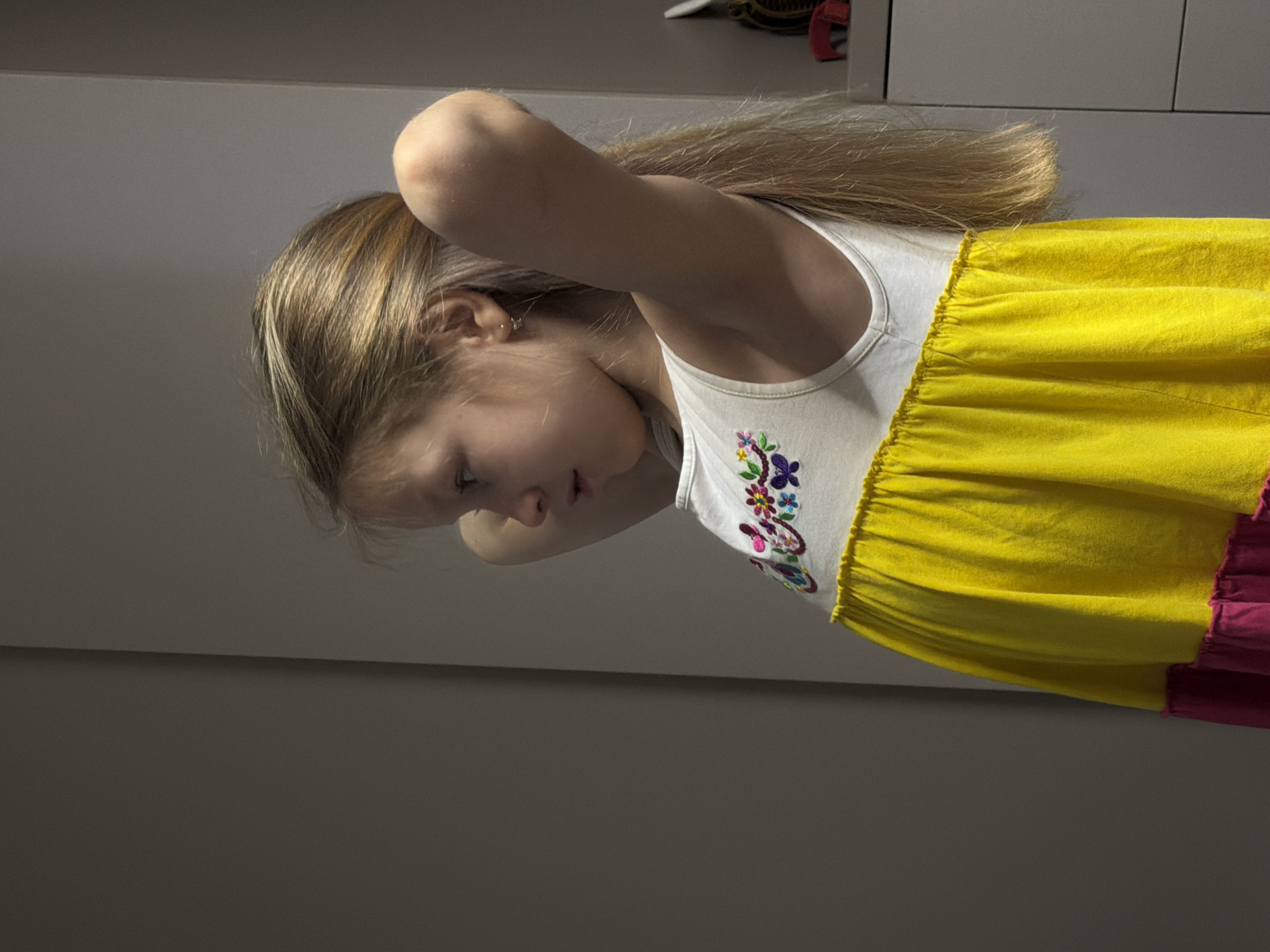 A young girl is seen at an angle, wearing a white top with floral embroidery and a yellow dress with a pink hem. She has light blond hair and her arm is raised near her head. The background consists of a gray wall and a cabinet with hair accessories on top. The location is specified as Dubovaya, Russia.
