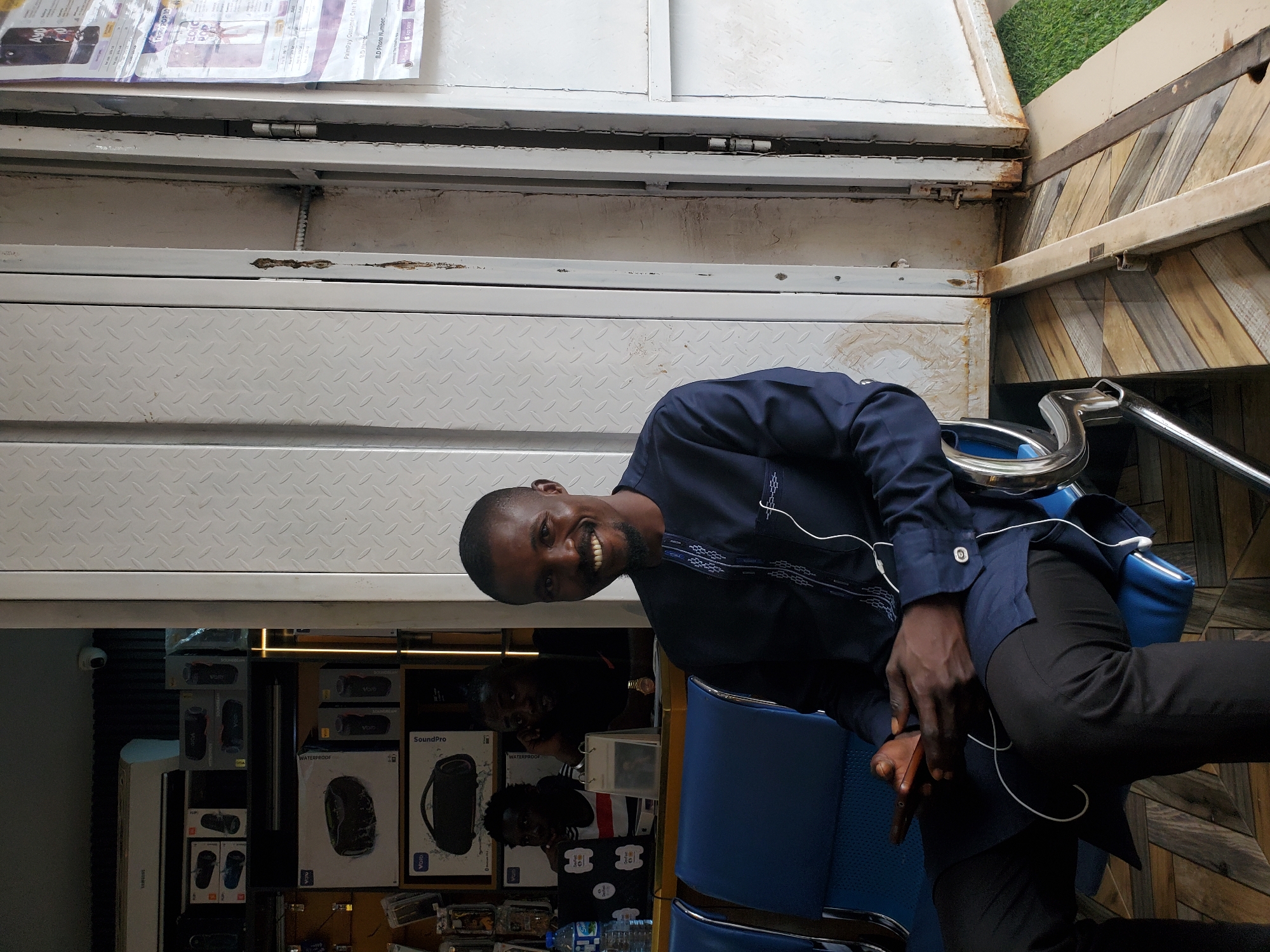 A man in a blue shirt and black pants is sitting in a blue chair. He is smiling and looking at the camera. The background shows shelves stocked with various electronic devices, including speakers and headphones, in their packaging. Some of these packages have text such as "SoundPro" and "WATERPROOF". There is also a sign visible with text including "PalmPay Customer Care Te" and "B.D Phone Number". The setting appears to be an electronics store. The lighting suggests it is daytime, and there are no obvious weather cues. The man's expression conveys a positive and friendly emotion.