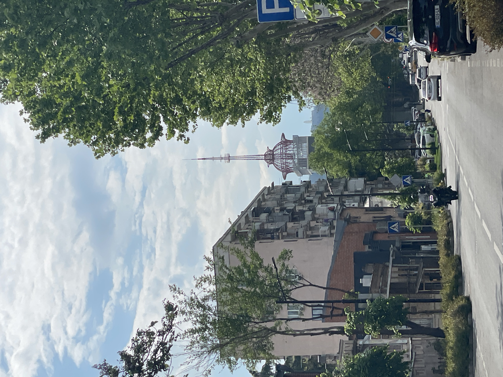 The image depicts a street scene in Tbilisi, Georgia, with the Tbilisi TV Broadcasting Tower visible in the distance. The view is slightly obscured by trees and buildings. A street with cars and pedestrian crossing signs is visible on the right. The architecture is mixed, featuring buildings with numerous balconies. The sky is partly cloudy.