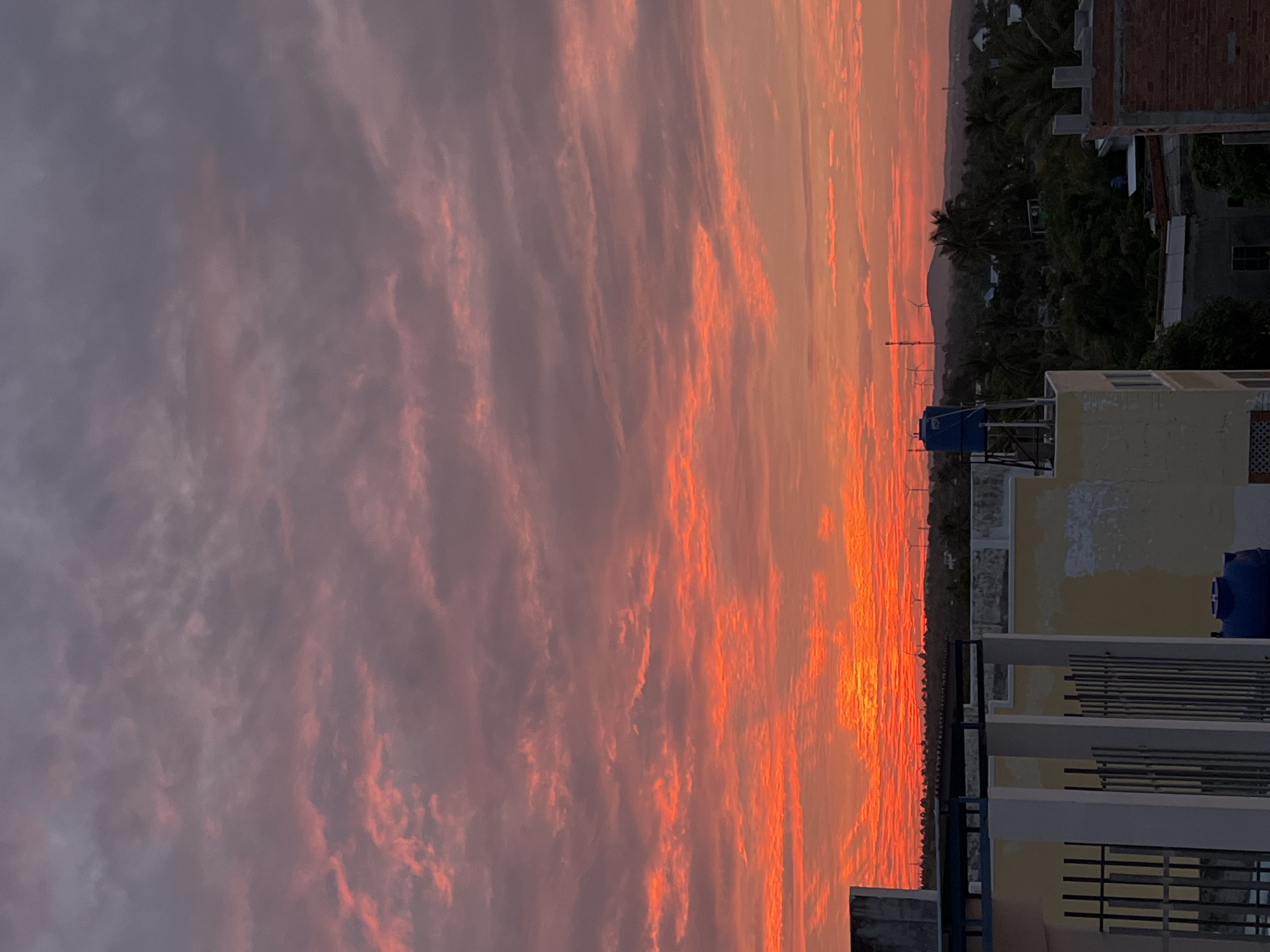 This is a photograph of a sunset over Mũi Né, Vietnam. The sky is filled with vibrant orange and pink clouds, a common sight during sunset in the region. In the foreground, the rooftops of several buildings are visible, constructed with concrete and painted in various shades of white and yellow. Some buildings feature metal railings and decorative elements. A blue water tank is perched on top of one of the buildings, indicating typical rooftop infrastructure. In the middle ground, a dense green forest stretches across the landscape, punctuated by palm trees. Beyond the trees, a distant hill is visible, topped with what appear to be wind turbines. The overall scene depicts a coastal urban environment during a colorful sunset.