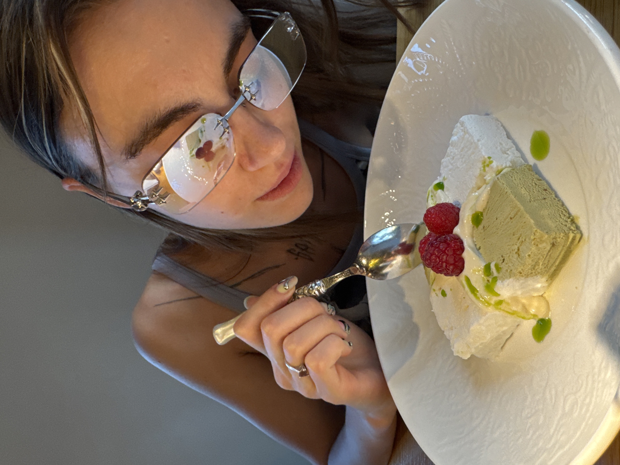 A close-up, high-angle shot captures a person, likely a woman, looking down at an artfully presented dessert, seemingly preparing to eat it in an indoor setting in Moscow, Russia.
The subject has long, dark hair with lighter streaks, partially obscuring her face from this angle. She wears distinct rimless glasses with decorative silver-toned elements on the sides; the lenses reflect light and subtle floral patterns. Her gaze is directed downwards, focused on the food. She holds an ornate, silver-colored spoon in her right hand, which displays light-colored nails, some with designs, and a silver ring on one finger. A blurred tattoo is visible on her chest/shoulder area.
The dessert is served on a large, textured white plate. It features a prominent piece of white, airy meringue or a similar light dessert, accompanied by a rectangular block of green-hued ice cream or mousse, possibly matcha-flavored. Two fresh red raspberries and a vibrant green sauce, drizzled artistically, complete the presentation.
The image emphasizes the dessert and the woman's interaction with it, with a softly blurred background hinting at a table or an indistinct indoor environment. There is no legible text visible in the image.