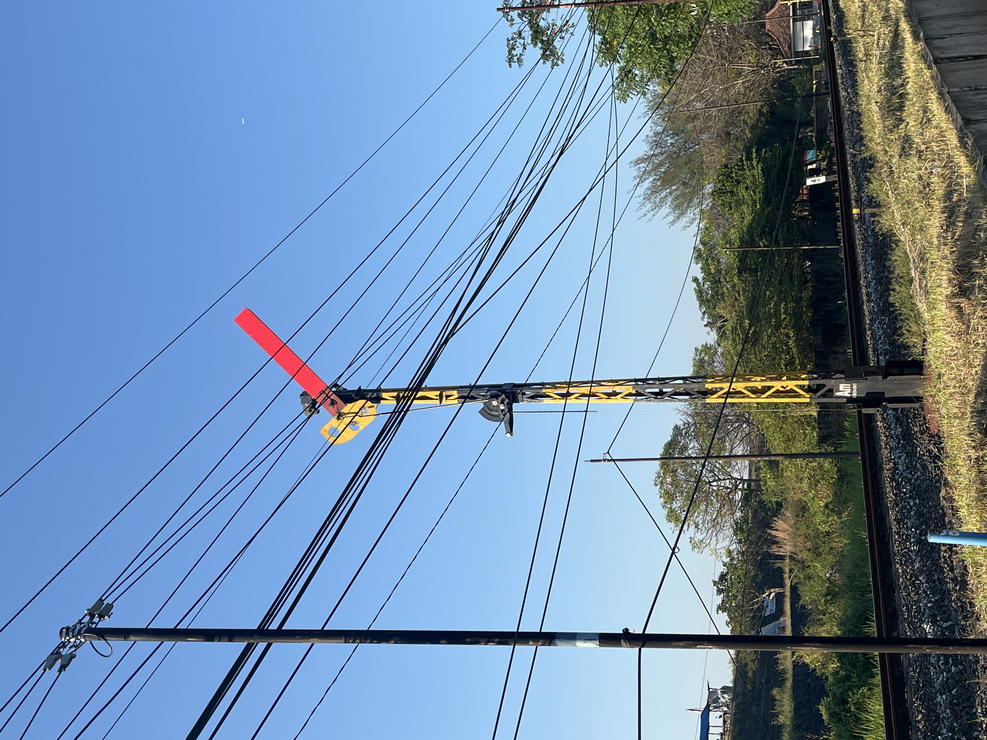 This is a full-frame, eye-level, outdoor, daytime, vertical photograph taken in Siring, Indonesia. The image features a railway signal with a red arm extended horizontally, indicating a stop or caution. The signal is mounted on a tall, black and yellow striped pole. Several dark electrical wires run across the frame, supported by a dark wooden pole on the left and another pole further to the right. The background shows a clear blue sky with a faint white object, possibly an airplane or distant cloud, visible in the upper right quadrant. Below the wires and signal, there are railway tracks, lush green vegetation, and trees. A concrete barrier runs along the bottom right of the frame. The scene appears to be in a rural or semi-rural area, with the focus on the railway infrastructure. No people are visible in the image.