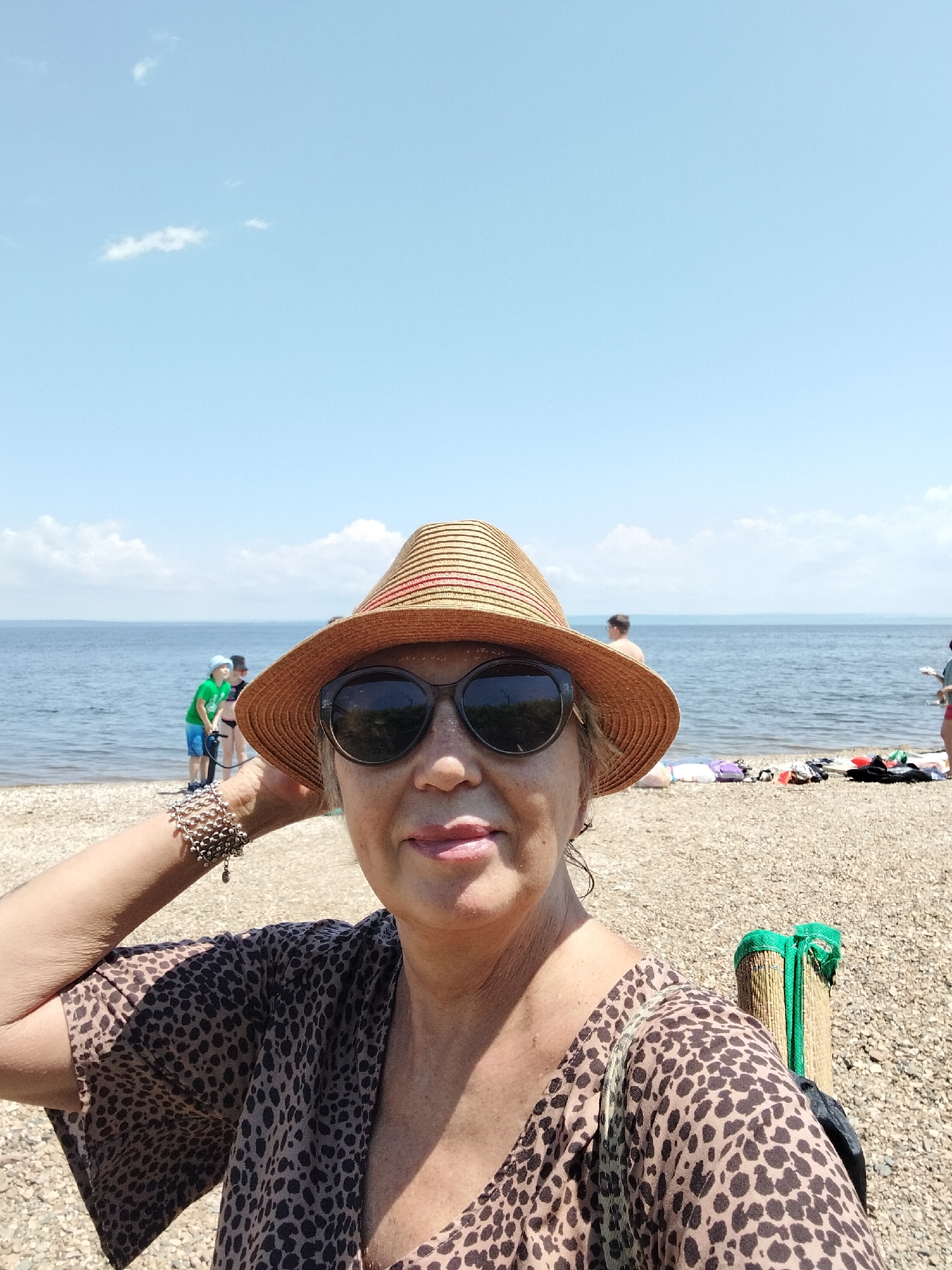 This is a selfie of a woman on a beach. She is wearing a wide-brimmed straw hat with red and brown stripes, large dark sunglasses, and a leopard-print top with flutter sleeves. She has a multi-strand silver bracelet on her left arm. Her right arm is raised, and she appears to be adjusting her hat or shielding her face from the sun. The background shows a body of water, likely the sea, under a clear blue sky with a few wispy clouds. On the beach, there are other people in the distance, some in swimwear, and beach items like towels and bags. The beach itself is covered in small pebbles. The scene is consistent with a sunny day at a coastal resort, potentially on the coast of Vladivostok, Russia, given the prevalence of such environments in the region.
