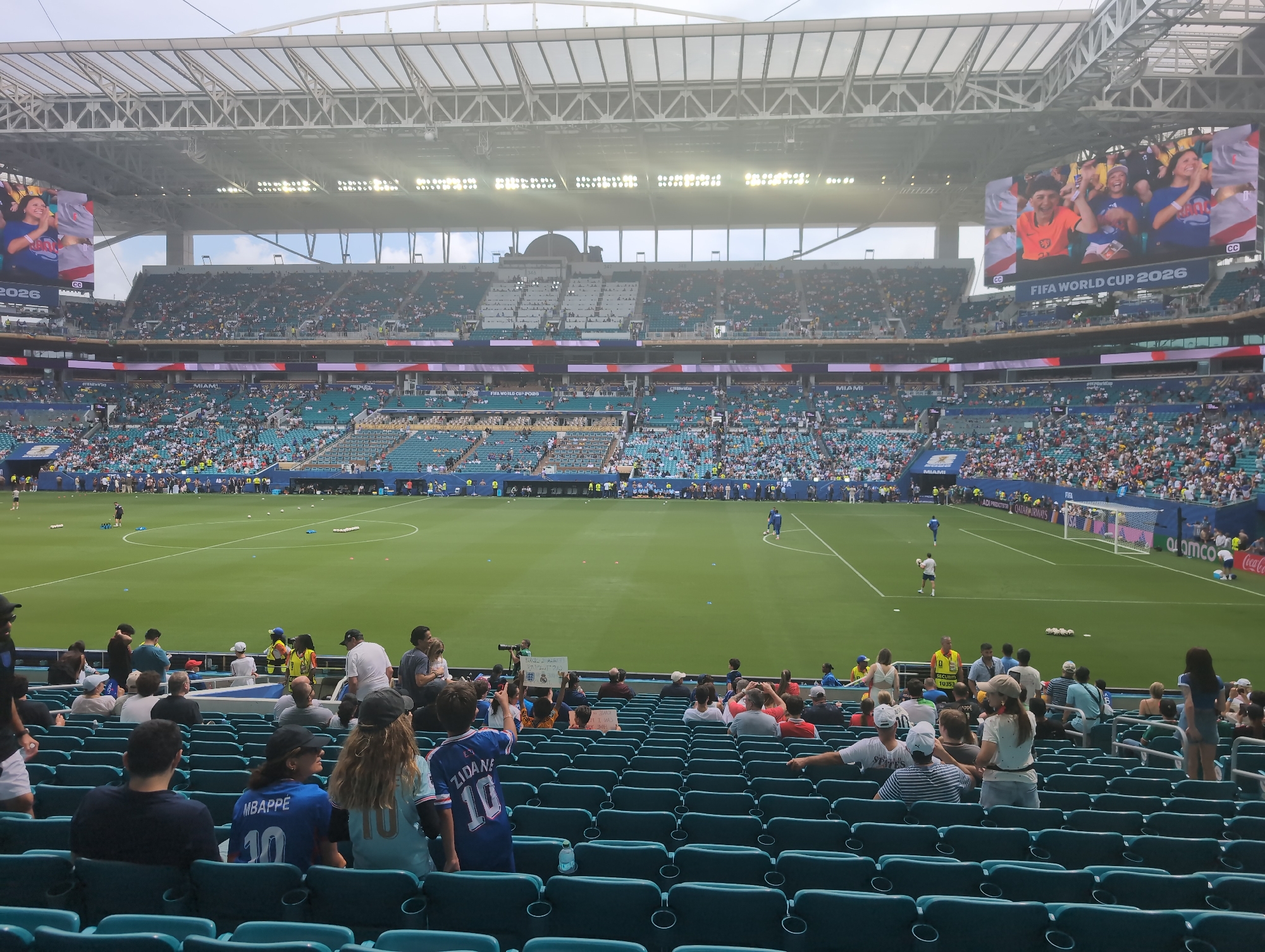 This image captures a wide view of the Hard Rock Stadium in Miami Gardens, Florida, United States of America, during a soccer match. The stadium is filled with spectators seated in tiered green seats, many of whom are wearing team jerseys. The field is a vibrant green, marked with white lines, and several soccer balls are visible on the grass. In the background, a large screen displays a montage of excited fans, with the text "FIFA WORLD CUP 2026" prominently featured. The stadium's retractable roof is partially open, allowing natural light to illuminate the scene. A diverse crowd of people, varying in age and gender, are present, with some standing and others seated, engaged in watching the game. Security personnel in yellow vests are also visible, ensuring the safety of the event. The overall atmosphere suggests a lively and energetic sporting event.