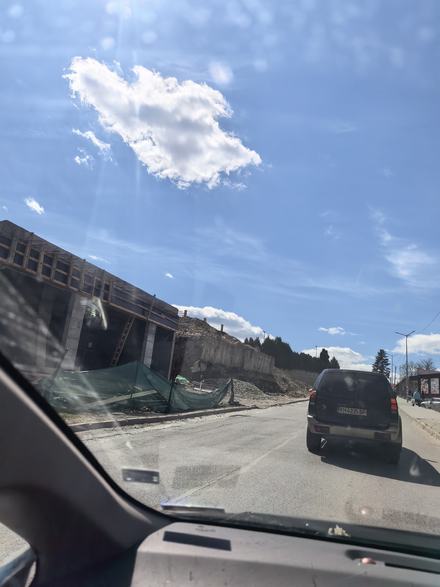 The image depicts a view from inside a car, looking out onto a street in an urban area. A dark-colored SUV is driving ahead, with a license plate visible that reads "KH 6235 BP." The road is asphalt, showing signs of wear and tear.
To the left of the road, there appears to be construction underway. A concrete structure with wooden framework is visible, partially obscured by green construction netting. A pile of dirt and debris sits nearby. In the background, trees can be seen on a slope.
On the right side of the road, there is a sidewalk with streetlights and what appears to be a bus stop or small kiosk. Two people can be seen in the distance on the sidewalk.
The sky is blue with scattered clouds. The image seems to be taken on a sunny day, based on the lighting and shadows.