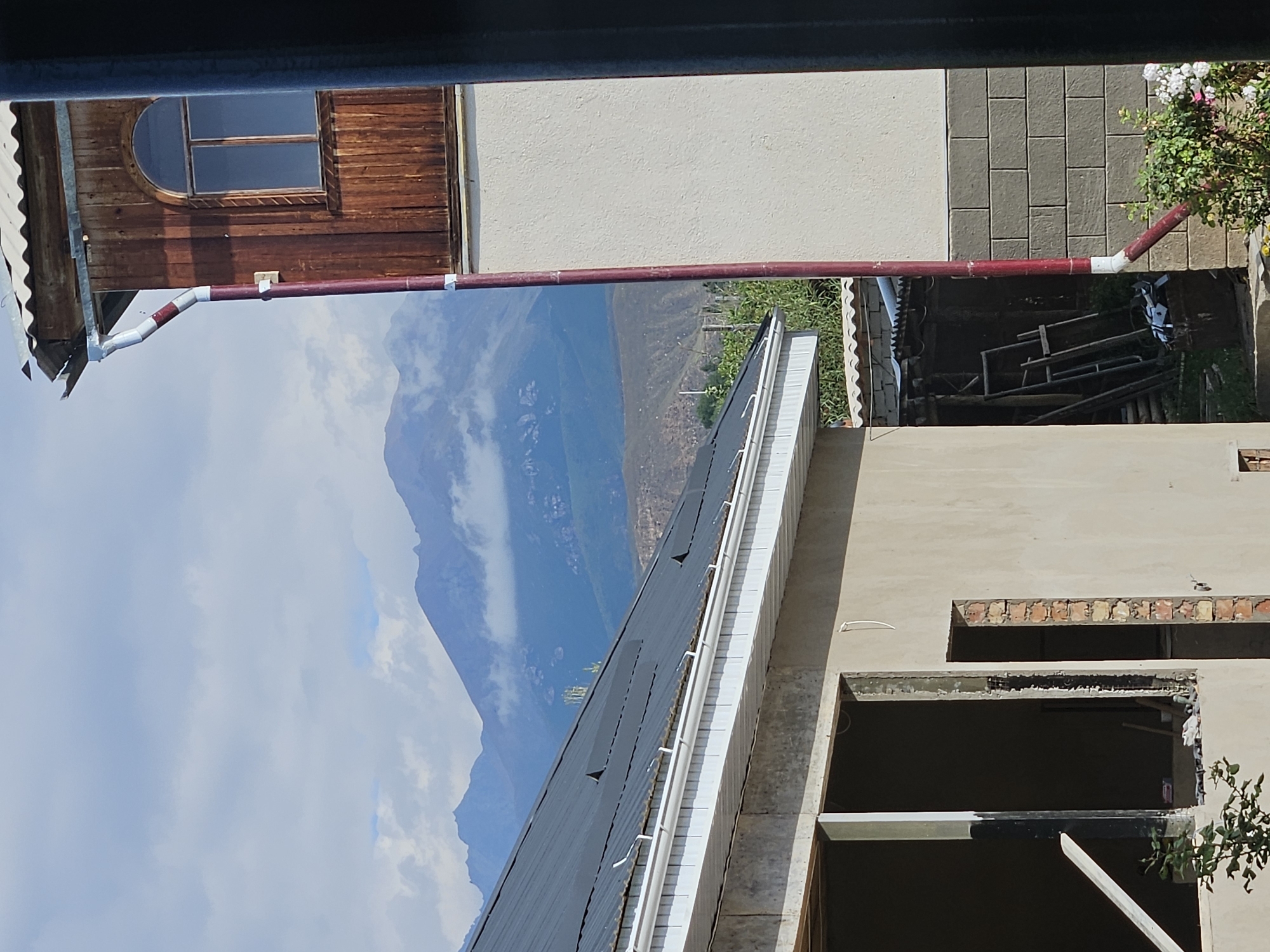 This image captures a view from a window looking out onto a rural or semi-rural landscape in Bulan-Segettu, Kyrgyzstan. In the foreground, construction is underway on a building with a gray concrete foundation and walls. A dark gray metal roof is visible, with a white gutter system installed along its edge. To the right, a two-story structure features a second-floor balcony with wooden siding and a arched window. A red downspout runs vertically along this building. Behind the immediate structures, a sloping, arid hillside is visible, with sparse vegetation. The background is dominated by a range of rugged mountains partially obscured by dramatic, cloudy formations. The sky is overcast with diffused light. No people are visible in the image.