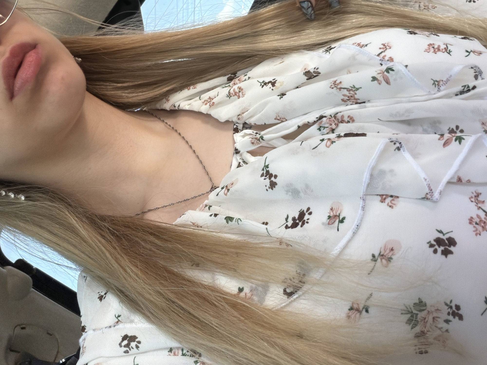 A close-up, angled shot shows a person, likely female, partially visible from the left side of the frame. Her lips are visible, colored with pink lipstick. She has long, blonde hair, with some strands falling across her face and shoulder. She is wearing a white floral print dress with ruffles at the neckline. A silver chain necklace is visible around her neck. The environment appears to be indoors, possibly in a car, as suggested by the partial view of a car interior and a glimpse of a blue sky with some white lines above through the window. The location in the city is Unknown, Unknown.
