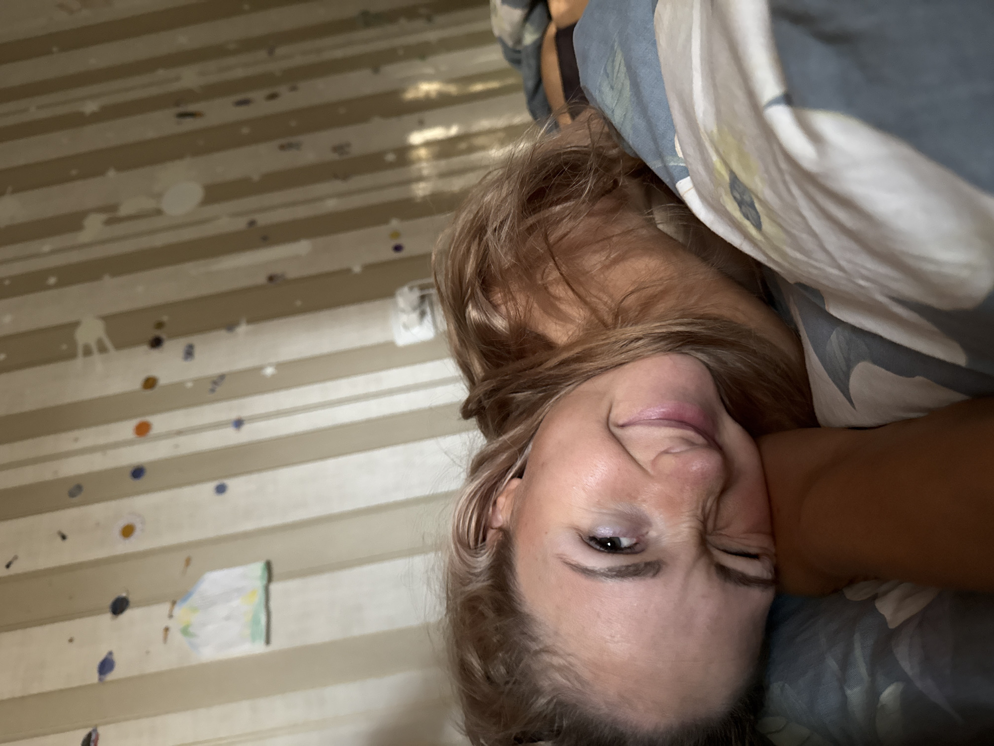 A selfie portrait depicts a light-skinned woman with light brown hair, partially covered by bed linens featuring floral and abstract patterns. She lies in bed, squinting her left eye, possibly winking. The background reveals a wall decorated with horizontal stripes and haphazardly placed colorful circular stickers, along with a child's drawing. The location is potentially inside a bedroom, most likely in Chaikovsky, Russia, although no definitive proof can be gleaned from the image itself. No specific actions or interactions beyond the selfie pose are visible.