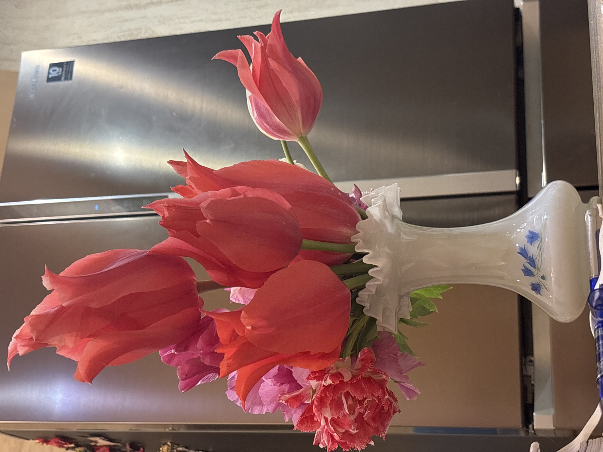 A bouquet of pink tulips and other pink flowers in a white vase with a ruffled neck and blue floral designs sits in front of a stainless steel refrigerator. The flowers are in various stages of bloom, with some fully open and others still partially closed. The background is out of focus. It is not possible to determine the location in Odesa, Ukraine from this image.