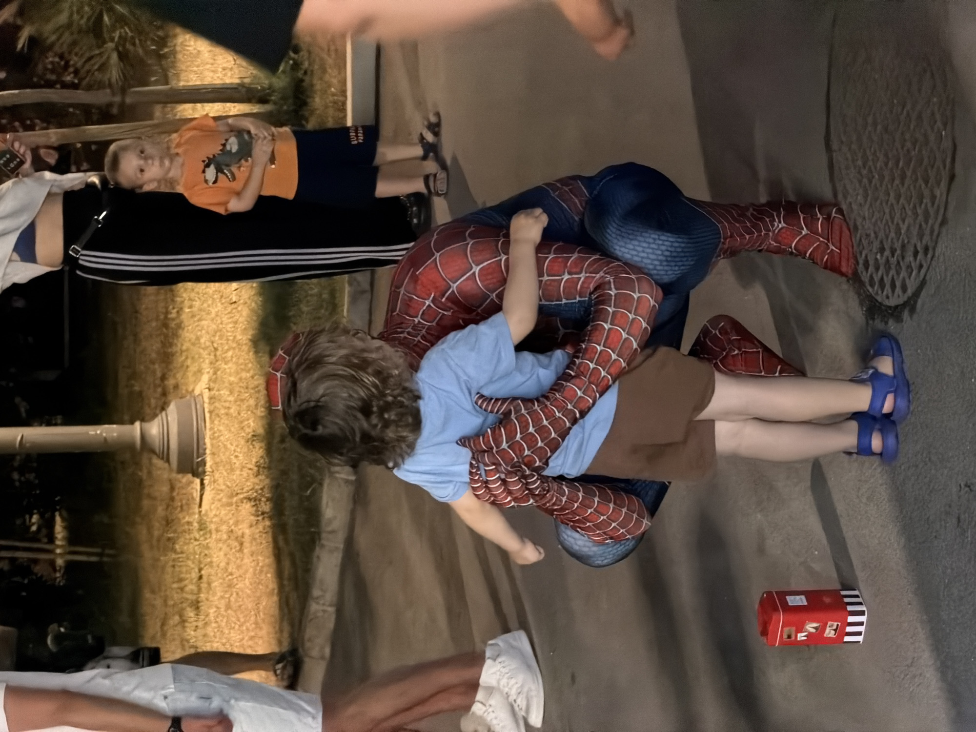 This media file shows a low-angle, sideways view of several people interacting outdoors at what appears to be dusk or evening.
In the foreground, a child dressed in a blue t-shirt and brown shorts is being held by a person in a full Spiderman costume. The Spiderman costume is red with blue accents and a black spiderweb pattern. The child's hands are resting on the Spiderman costume's chest, and they are looking towards the camera. The Spiderman character's arms are wrapped around the child. The child is wearing blue sandals.
To the left of the Spiderman costume, another child is lying down, wearing an orange t-shirt with a dinosaur graphic and dark blue shorts. This child is looking up towards the top of the frame. A person's legs are visible to the far left, wearing black pants with white stripes and white athletic shoes.
The background consists of a paved surface, a patch of dry grass, and a lamppost. A small red and white striped box, possibly a toy or container, is on the ground to the right. The lighting suggests it is either early morning or late evening. There is no visible text in the frame.