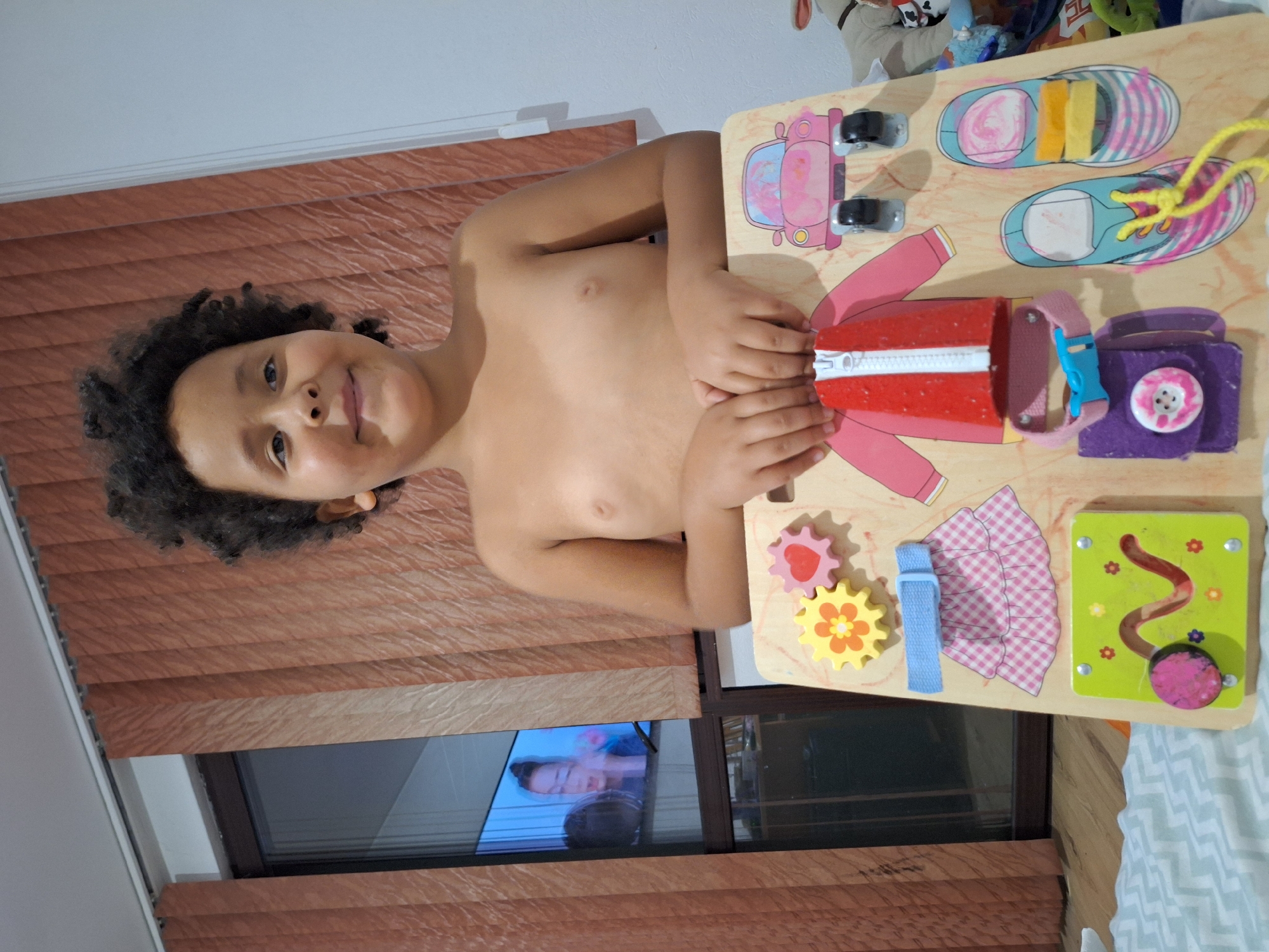 A child with curly dark hair and fair skin smiles at the camera, standing in front of a wooden activity board. The child is shirtless and has their hands resting on the activity board. The activity board features various attached objects, including gears, a pink and white checkered skirt with a blue belt, a jacket with a zipper, a pink collar with a blue buckle, a purple pouch with a button, a pink toy car with wheels, and two pairs of shoes with laces. The background includes vertical brown curtains and a window reflecting a screen showing a person. The scene appears to be indoors, likely in a residential setting. The city of Blagoevgrad, Bulgaria, is not visibly represented in this image.