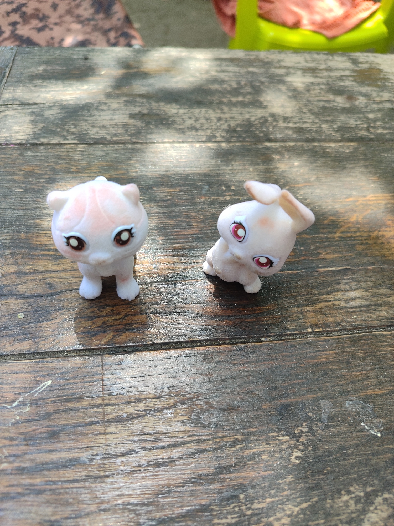 Two small, light-colored plastic figurines are positioned on a weathered wooden surface. The figurine on the left resembles a cat, with large, dark eyes featuring white highlights and delicate eyelashes. It has a rounded head with small ears and pink markings on its forehead and cheeks. Its body is white with subtle pinkish-orange shading. The figurine on the right appears to be a rabbit or similar creature, also with large, red eyes and white highlights. It has prominent, light-colored ears that are slightly bent and pink blush marks on its cheeks. The wooden surface is dark and shows signs of wear, with visible grain and some lighter patches. Sunlight casts dappled shadows across the scene. In the background, out of focus, is a bright lime green chair with a pink fabric draped over it. The setting is outdoors, suggested by the natural light and shadows.