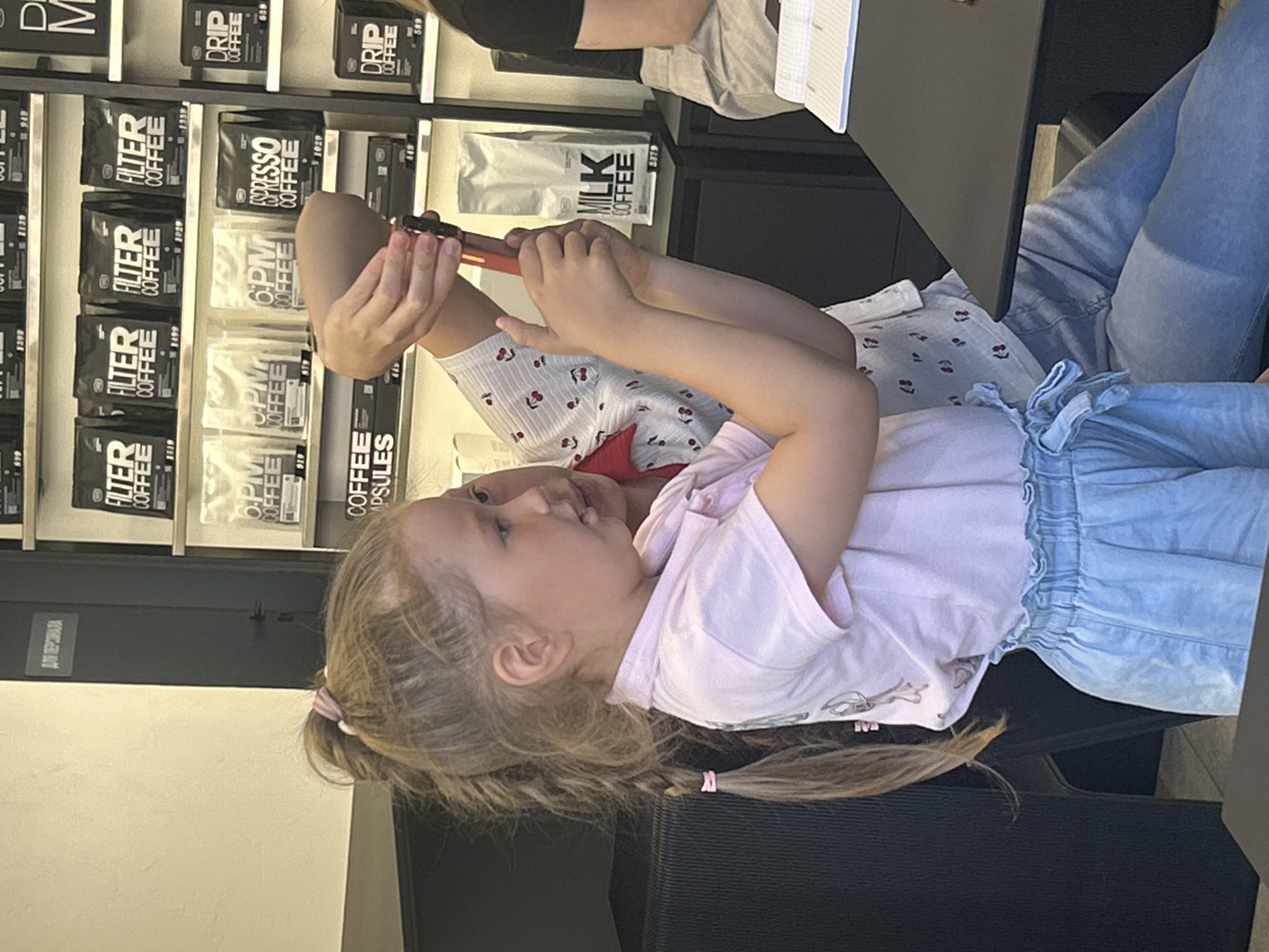 A young girl with blonde hair in a ponytail and wearing a pink t-shirt and light blue shorts is holding a red and black object. She is looking to her right. Behind her, an adult wearing a white shirt with red cherry prints and blue jeans is also holding the same object, assisting her. The background shows shelves stocked with bags of coffee, including brands like "FILTER COFFEE," "6 PM COFFEE," "ESPRESSO COFFEE," and "DRIP COFFEE," as well as a bag labeled "MILK COFFEE." The environment appears to be a cafe or coffee shop. The scene is likely in Izhevsk, Russia, based on the Cyrillic text visible on the sign in the upper left corner which reads "ДЛЯ ПЕРСОНАЛА" (FOR PERSONNEL).