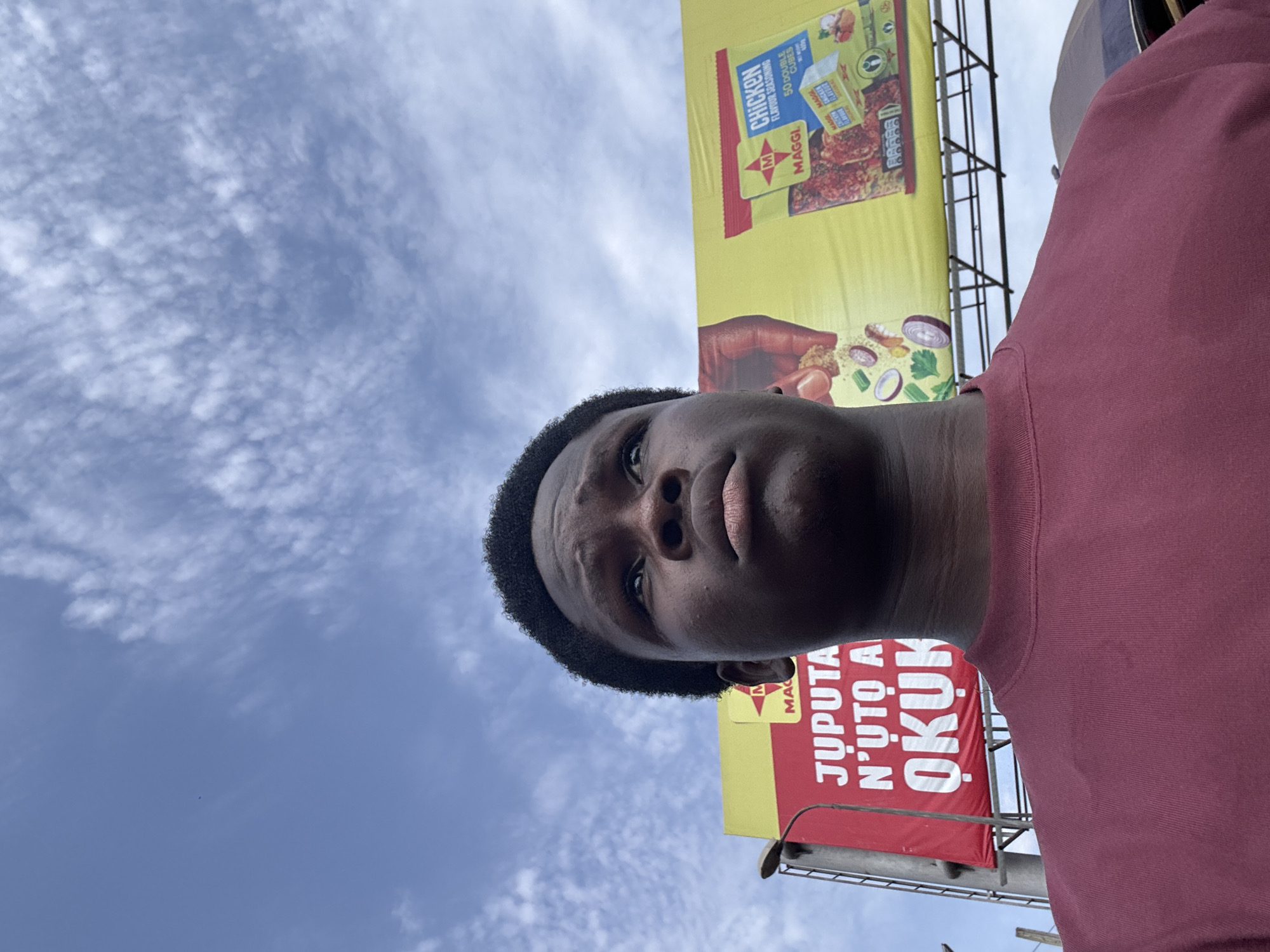 A low-angle, medium shot captures a young Black male in the foreground, looking upwards and to the right. He has dark, curly hair and is wearing a dark red, crew-neck t-shirt. Behind him, a large billboard is visible, featuring a red section on the left with white text that appears to be in an African language, and a yellow section on the right advertising Maggi chicken-flavored seasoning. The billboard is supported by a metal structure. In the background, a bright blue sky with scattered white clouds is present. The scene appears to be outdoors, possibly on a street in Onitsha, Nigeria, given the context of the Maggi advertisement, which is very popular in West Africa.
